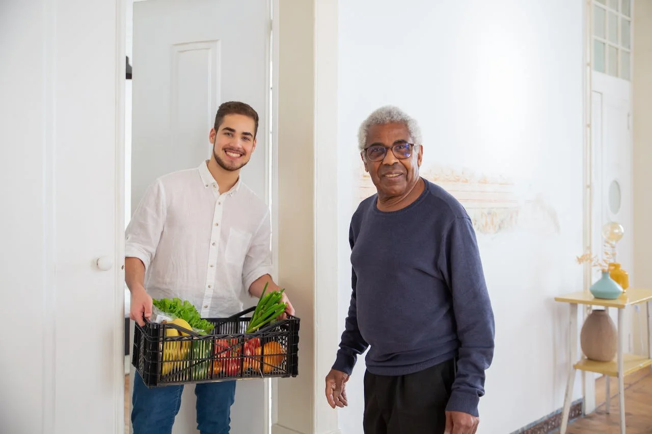 A young man delivering a basket of groceries to an elderly man indoors, both smiling.