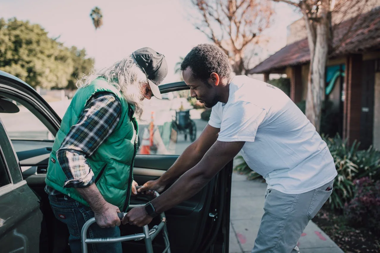 A young man helps an elderly woman with a walker get into a car outside a house. The woman is wearing a baseball cap, plaid shirt, and green vest. The young man is wearing a white t-shirt and gray pants. The scene is outdoors with trees and houses in the background.