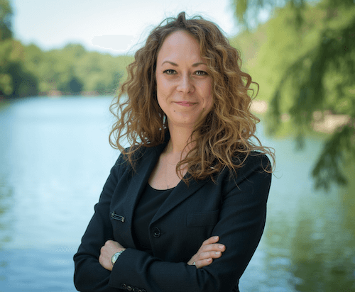 Frau mit lockigem Haar steht am Fluss im Freien, lächelt leicht, trägt schwarze Kleidung.