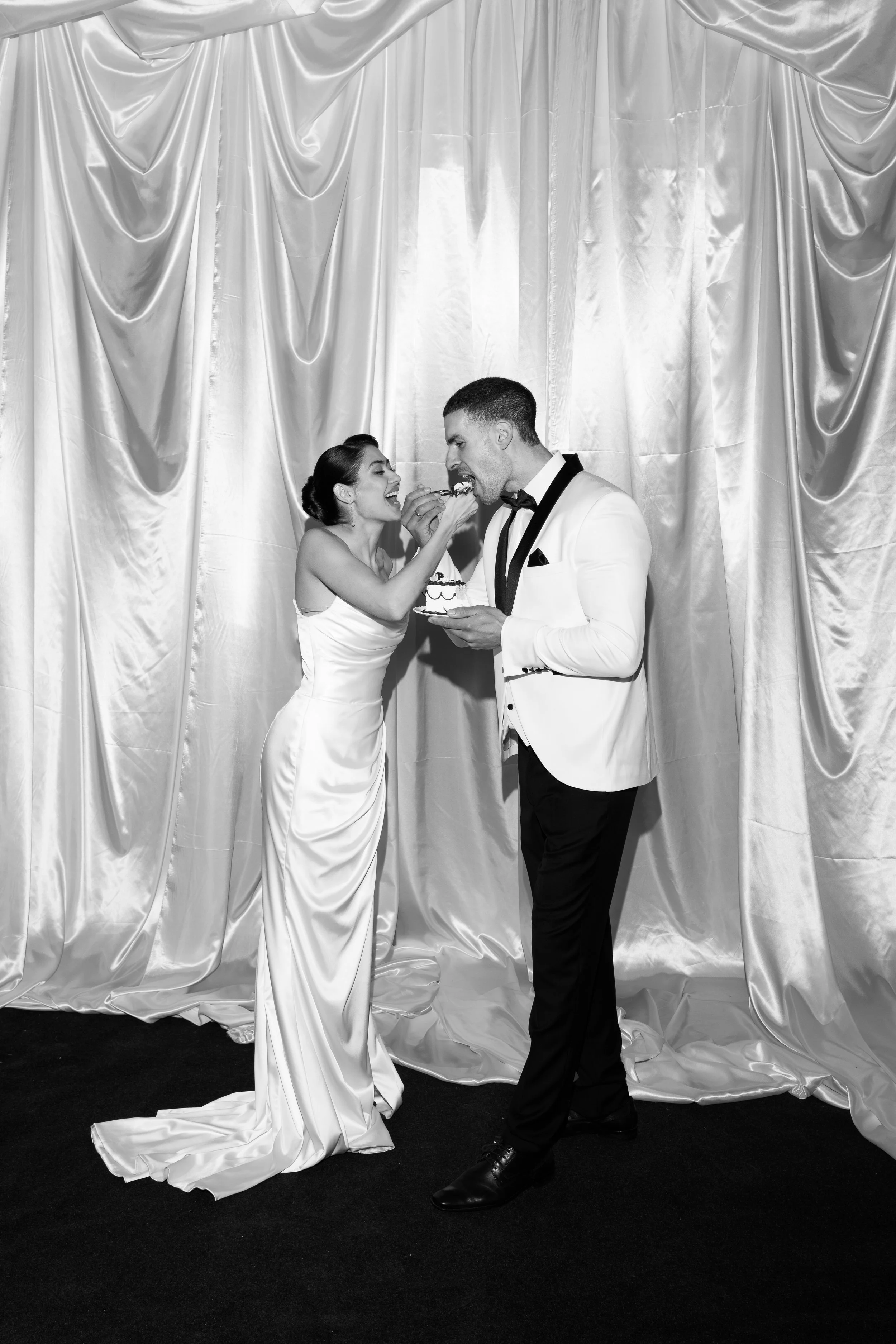 A bride and groom in formal attire sharing a piece of cake at their wedding reception, with a silky curtain backdrop.