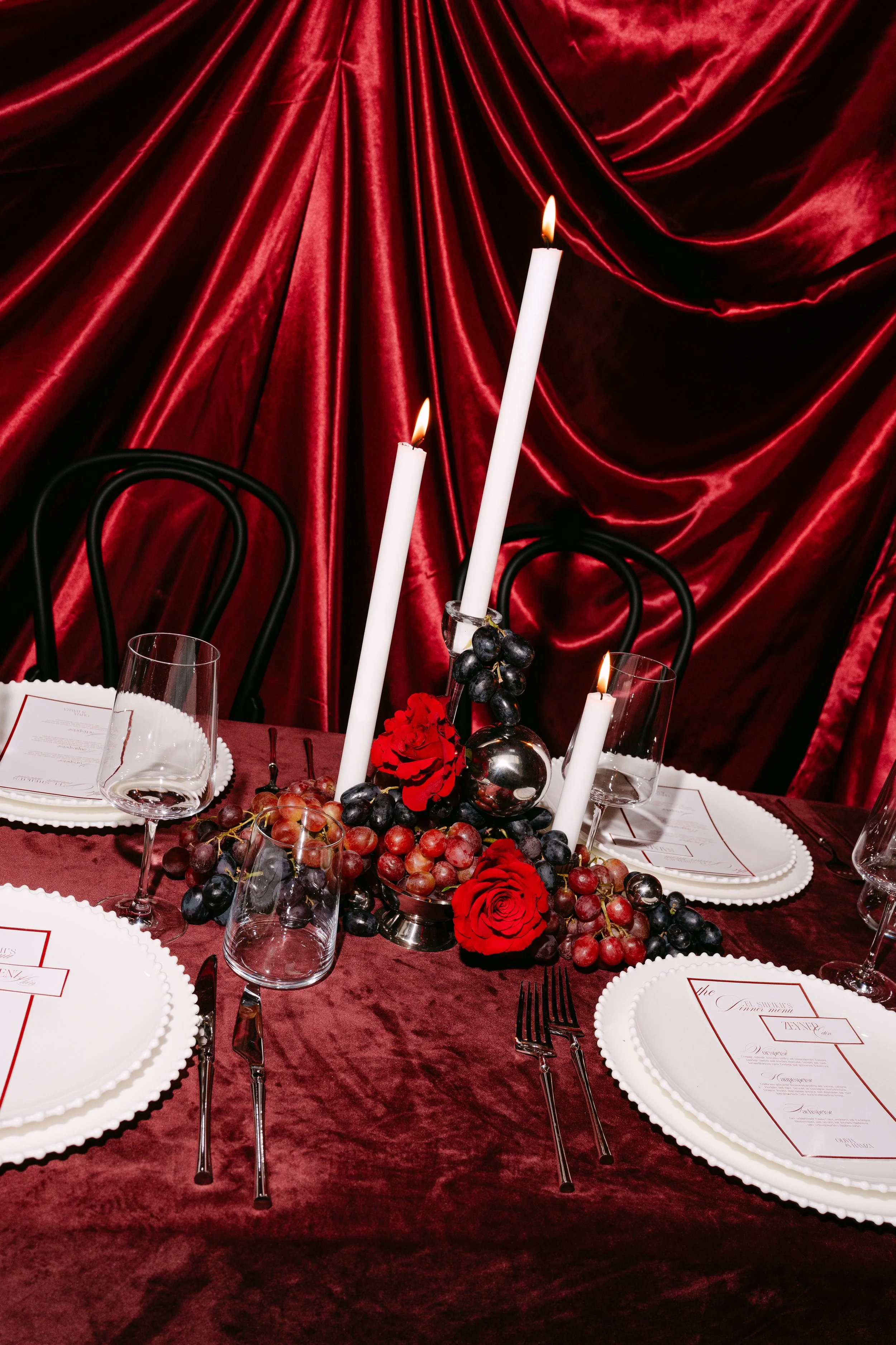 A table setting with white plates, wine glasses, and silverware on a burgundy velvet tablecloth. The centerpiece includes three tall white candles, red roses, black and red grapes, and silver decorative balls against a red velvet backdrop.