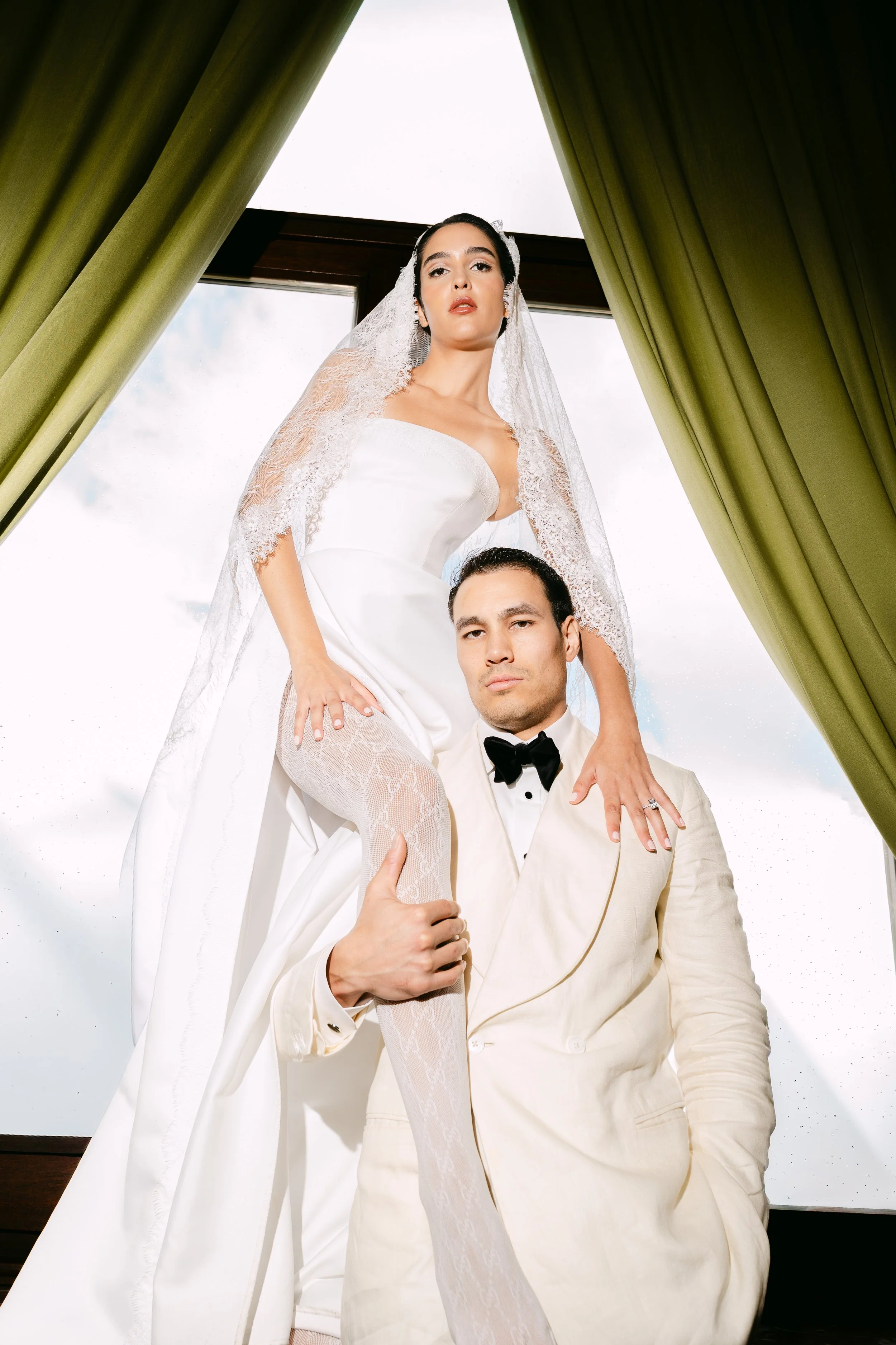 A woman in a wedding dress with a lace veil stands on a man's shoulders, in front of a window with green curtains. The man is dressed in a cream tuxedo with a black bow tie. Both are looking directly at the camera.