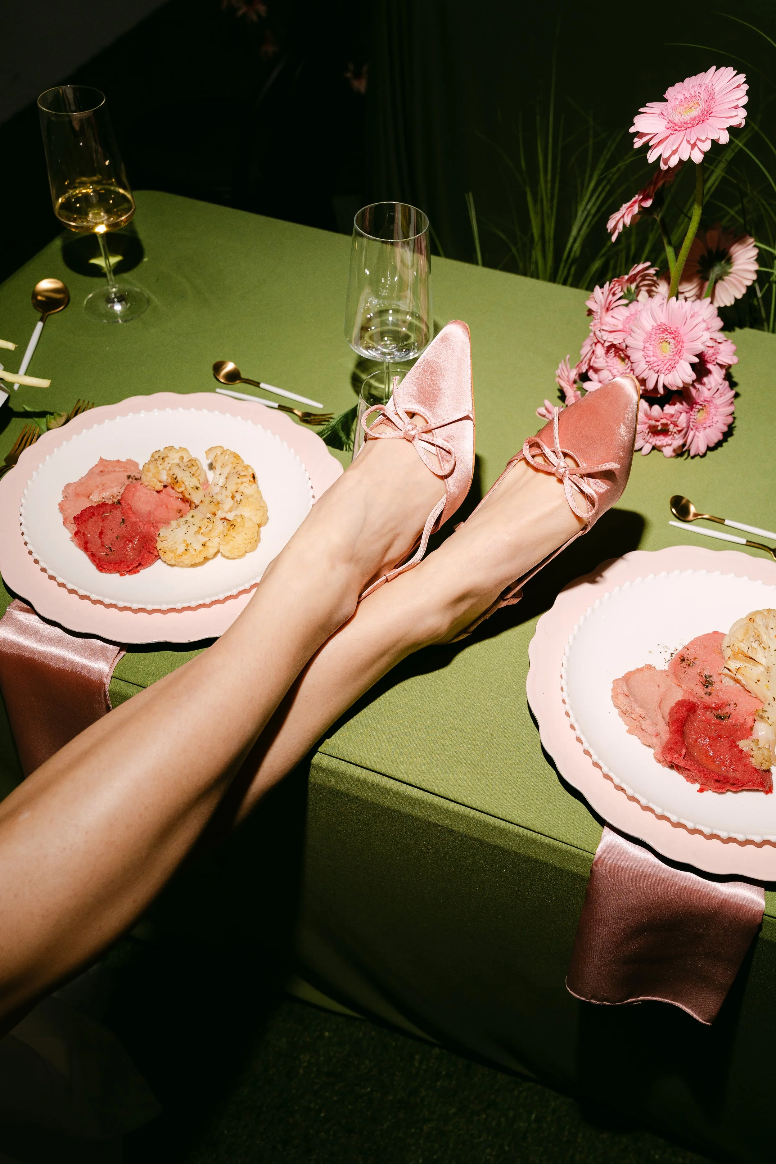 A table setting with pink shoes worn by a person reclining, pink flowers in a vase, empty plates, gold utensils, and champagne glasses on a green tablecloth with pink satin trim.