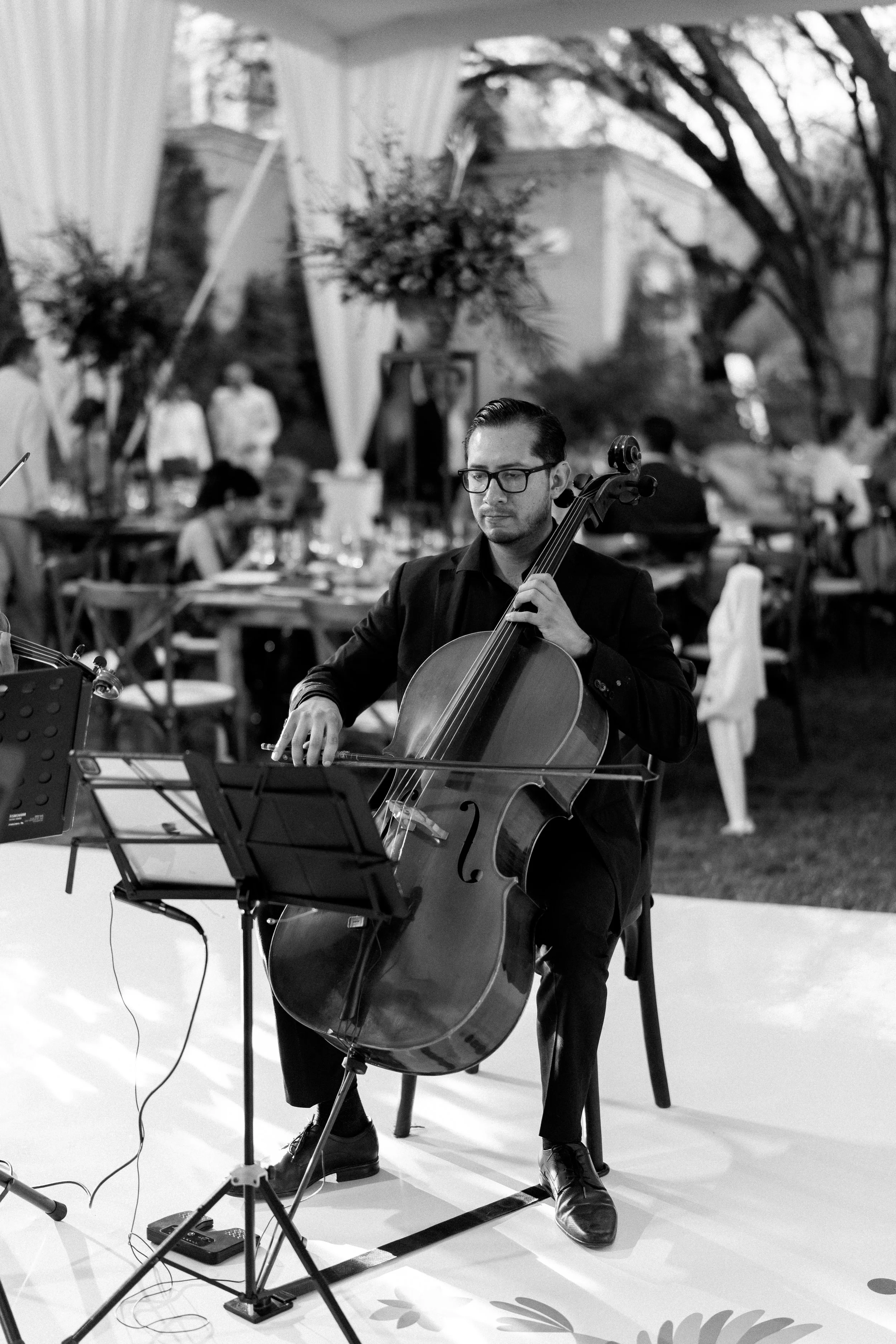A man playing a cello at an outdoor event, with tables and guests in the background.