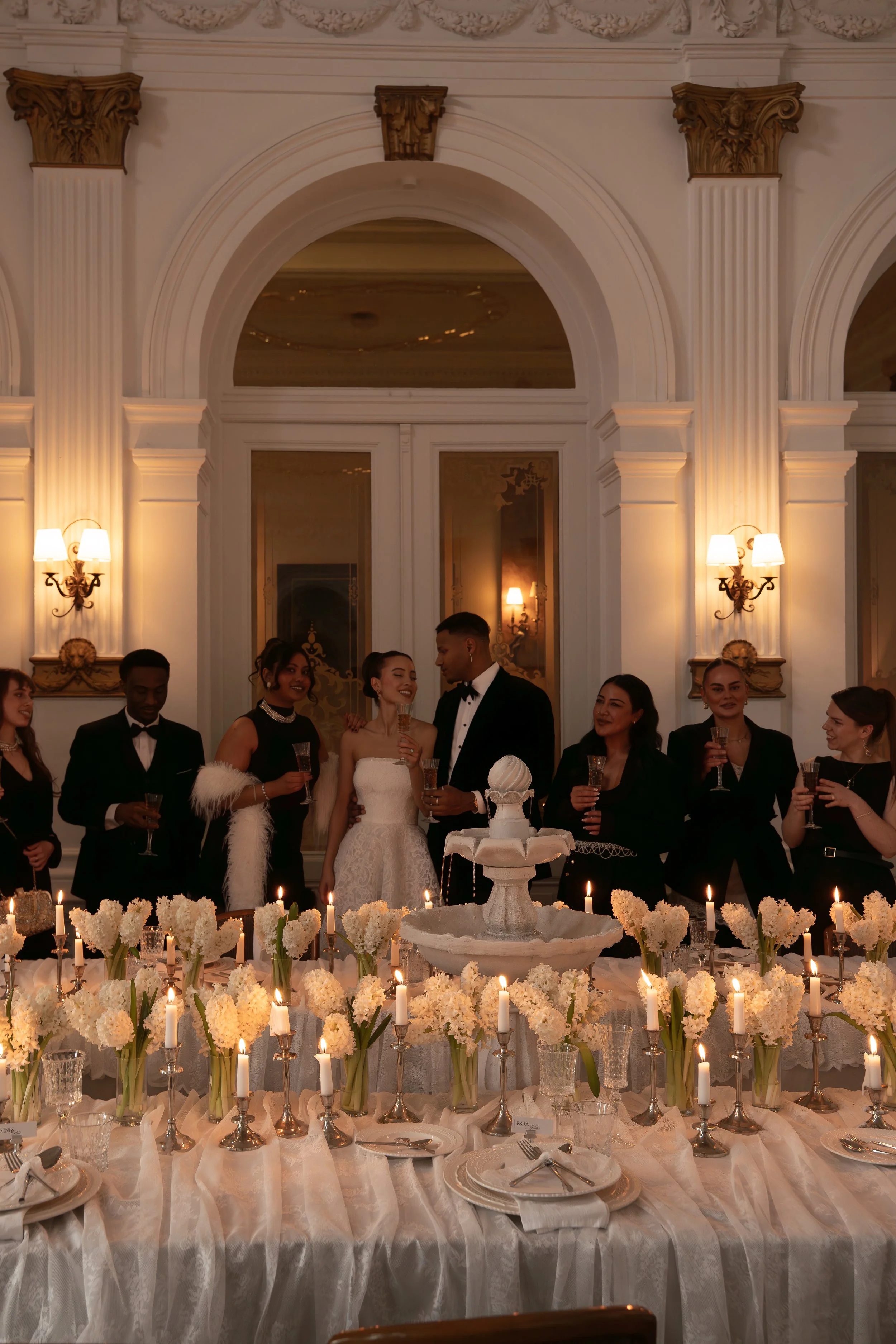 A bride and groom at a wedding reception cutting a tiered cake, surrounded by friends dressed in formal attire, in an elegant ornate ballroom with white walls, gold accents, and ambient lighting.