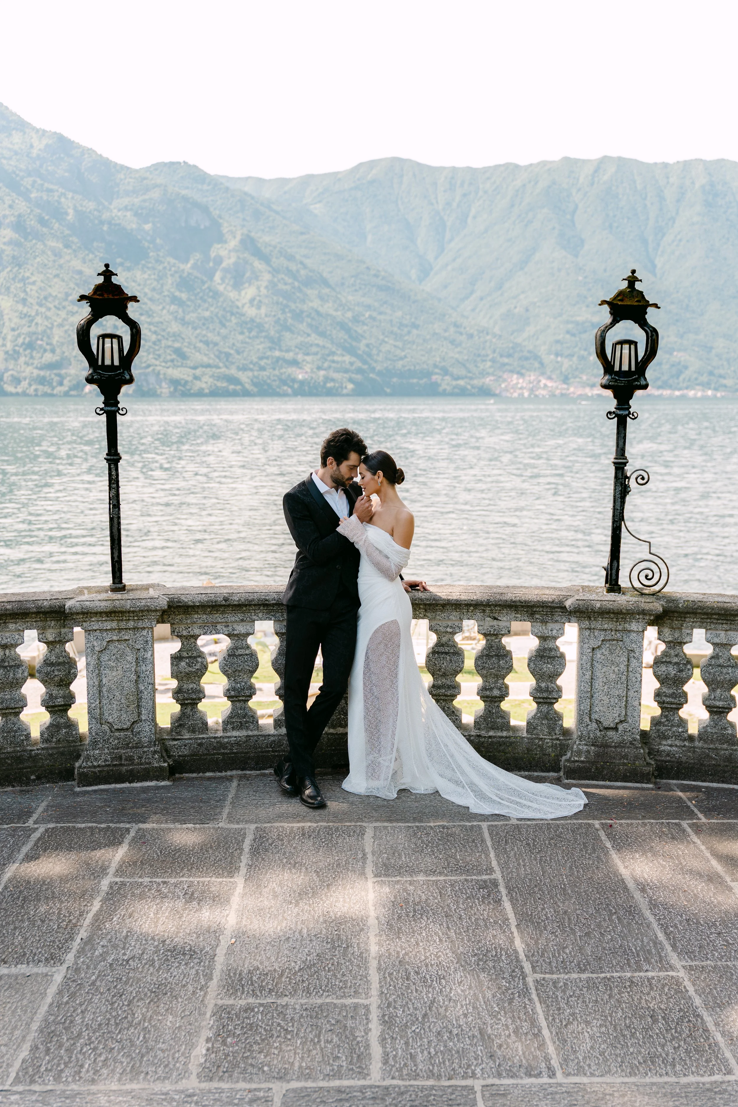 A bride and groom standing close together on a stone terrace by a lake with mountains in the background, during their wedding.