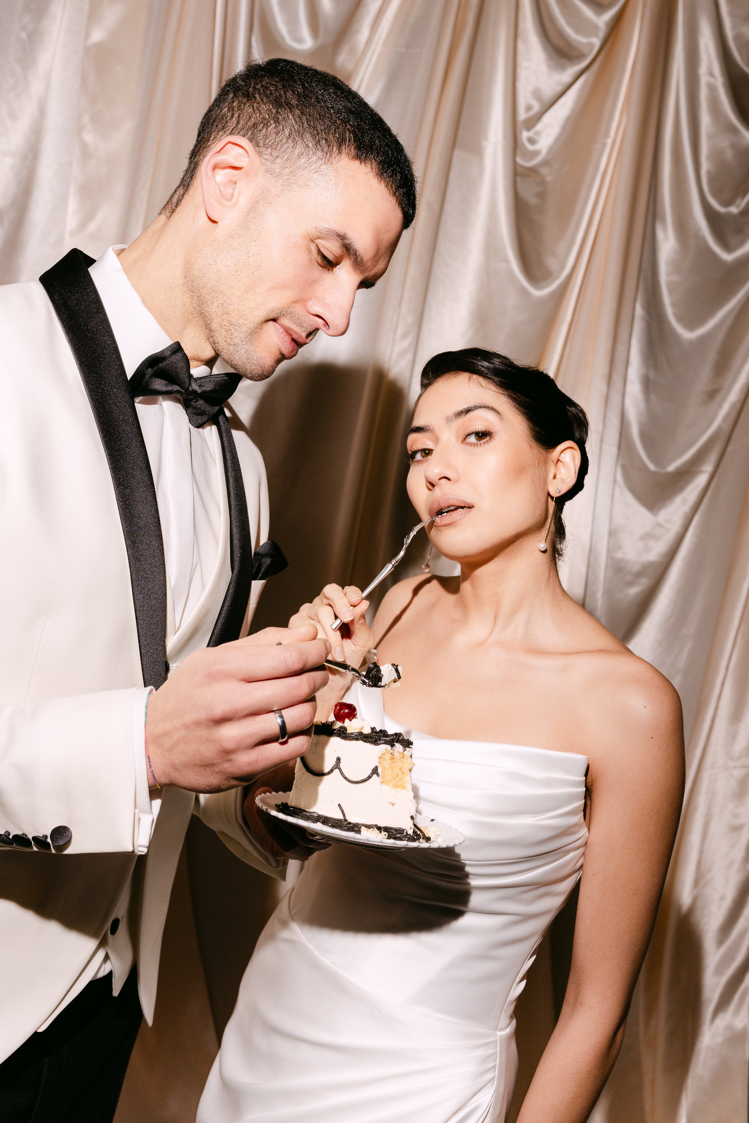 Bride and groom at a wedding, with the groom holding a slice of cake and the bride looking at the camera with a confident expression.