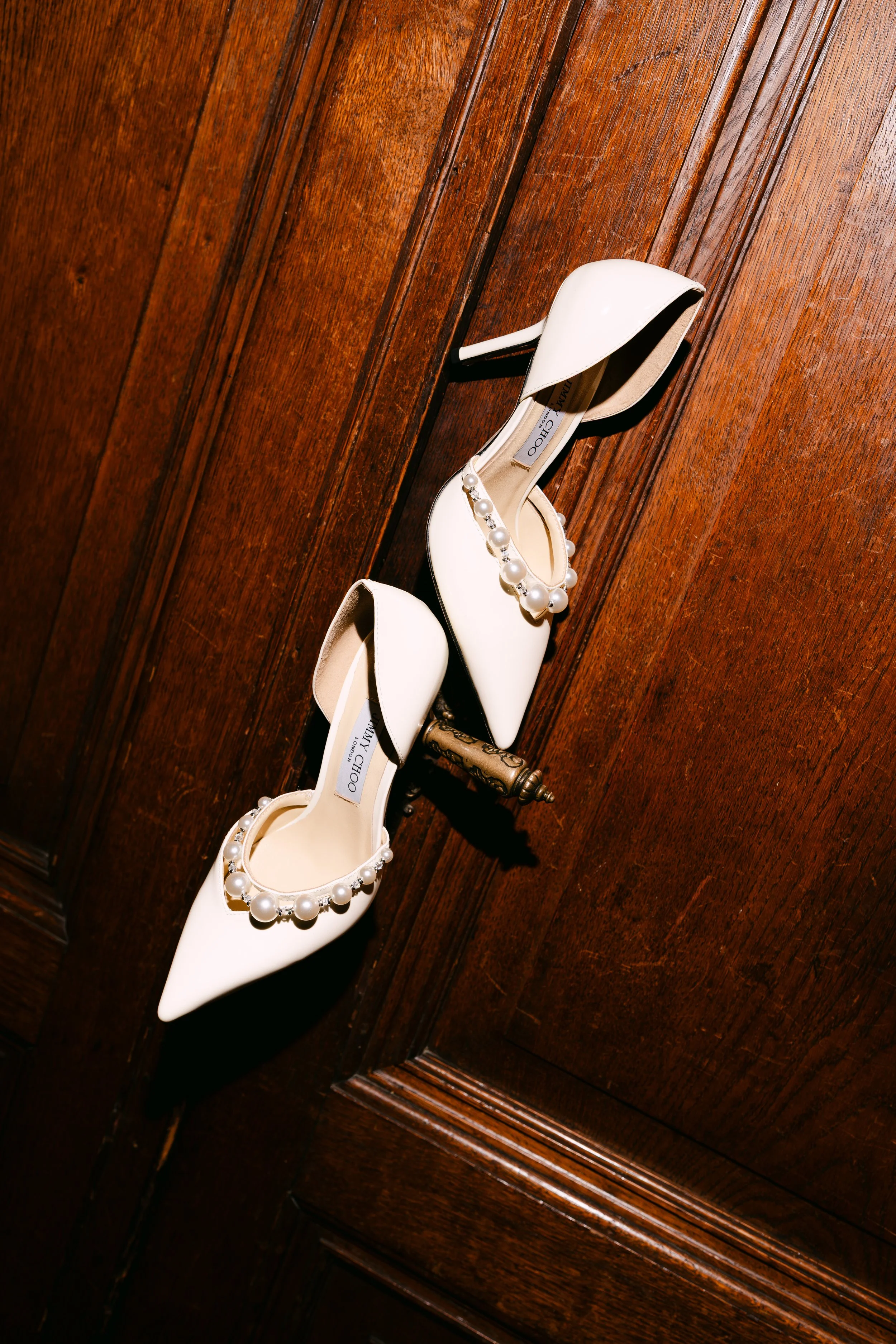A pair of white high-heeled shoes with pearl embellishments placed on a wooden surface.