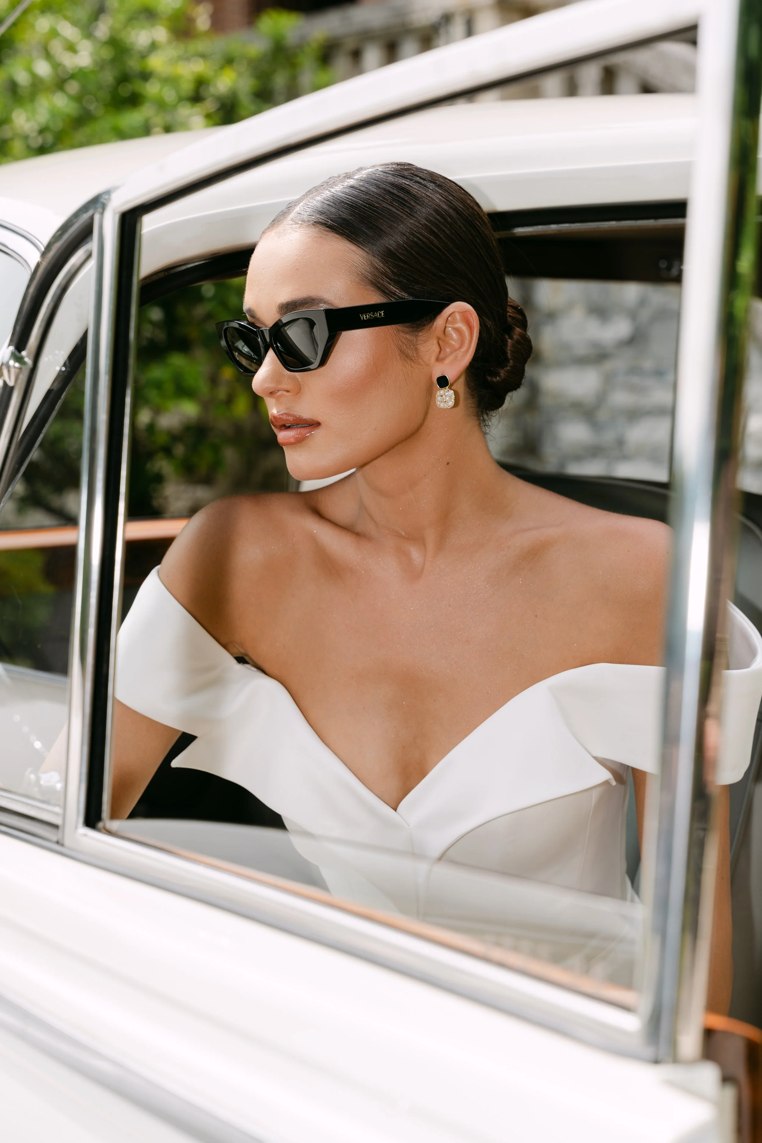 A woman in an elegant white off-shoulder dress with styled hair, wearing black Versace sunglasses and pearl earrings, seated in a classic white car.