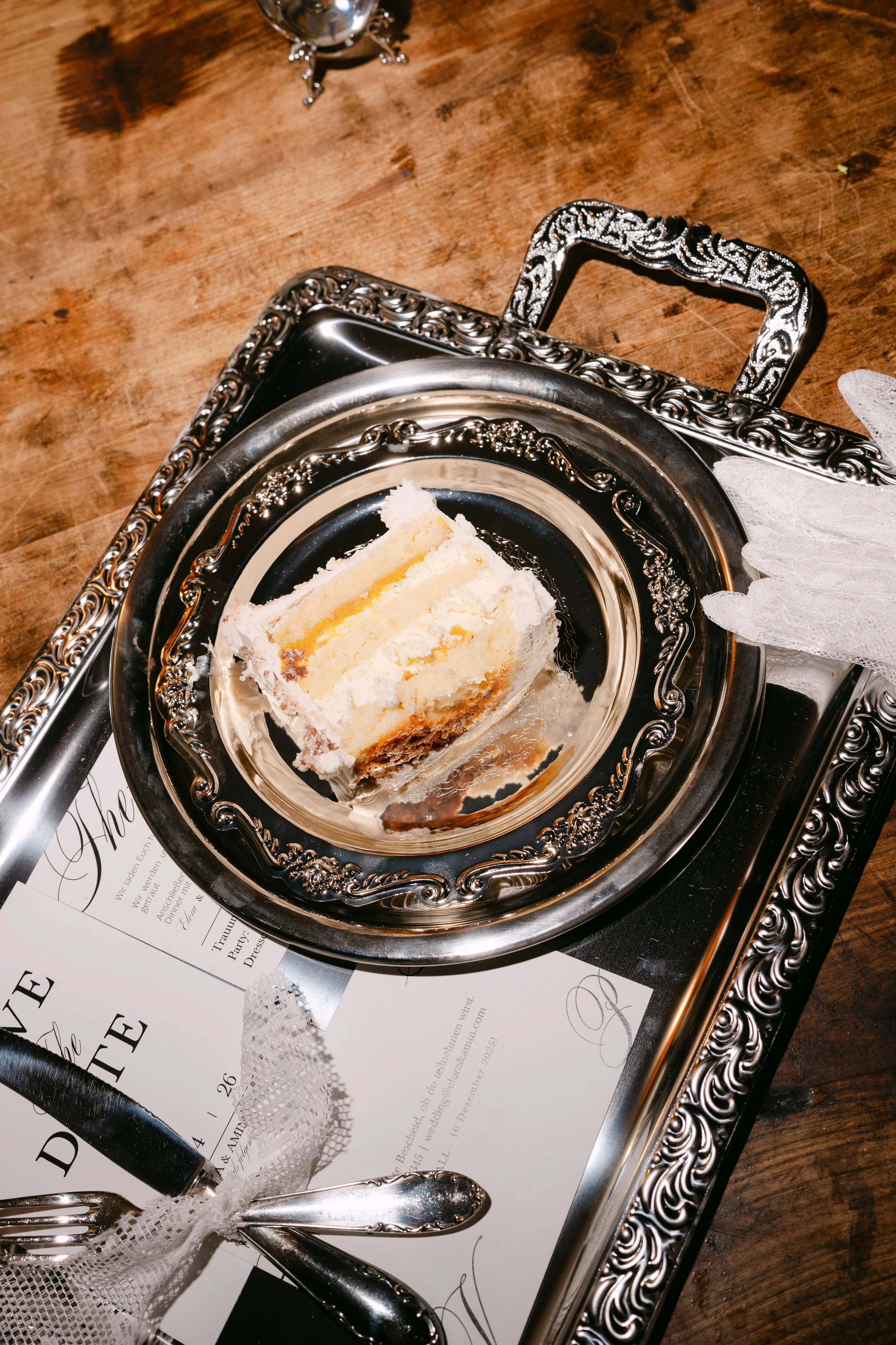 A slice of wedding cake on a silver ornate plate placed on a decorative silver tray, set on a wooden table, with a fork, a lace napkin, and wedding invitations nearby.