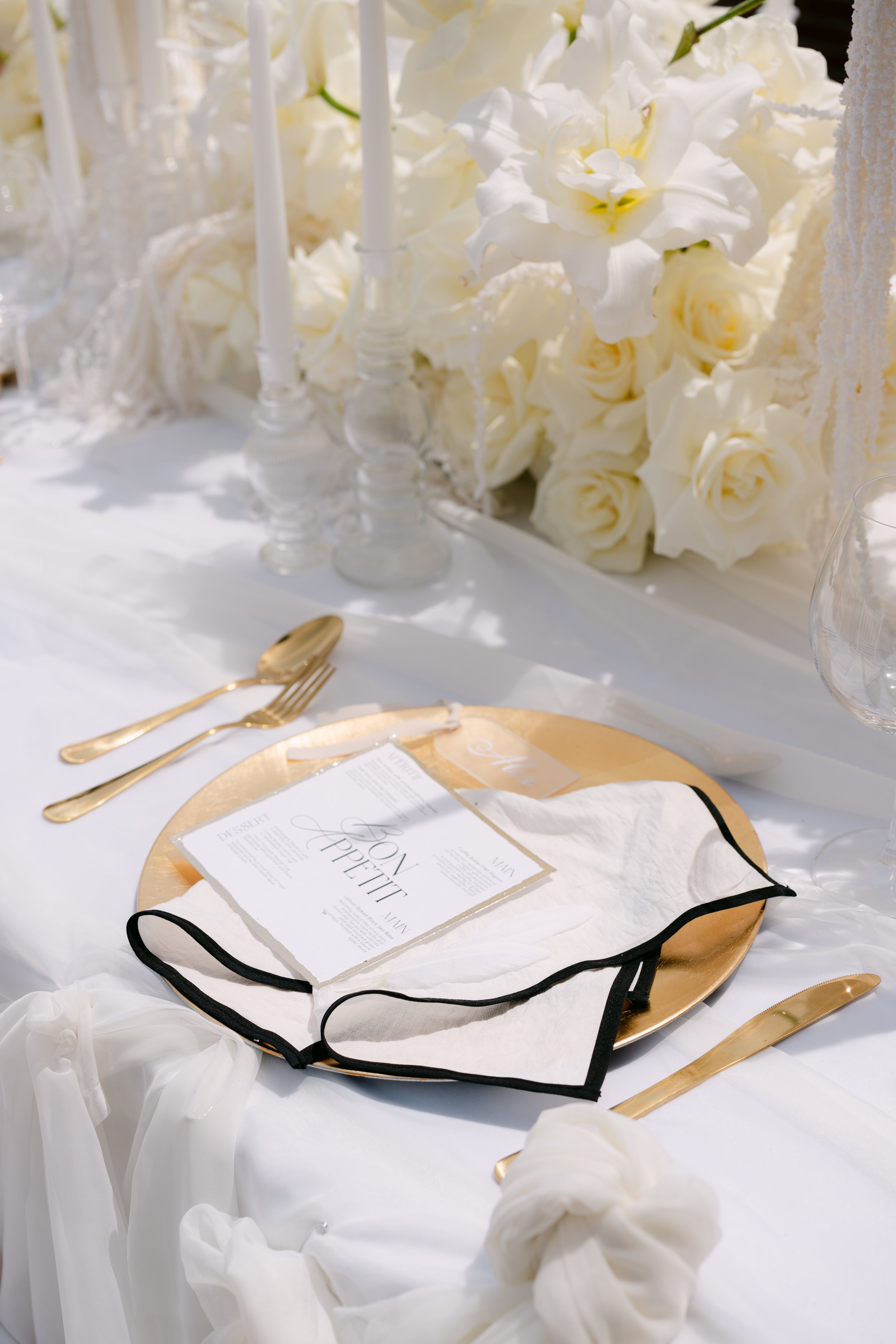 Elegant table setting with a gold charger, gold flatware, and white napkin with black trim, in front of a large floral arrangement of white roses and lilies, with candles in glass holders.