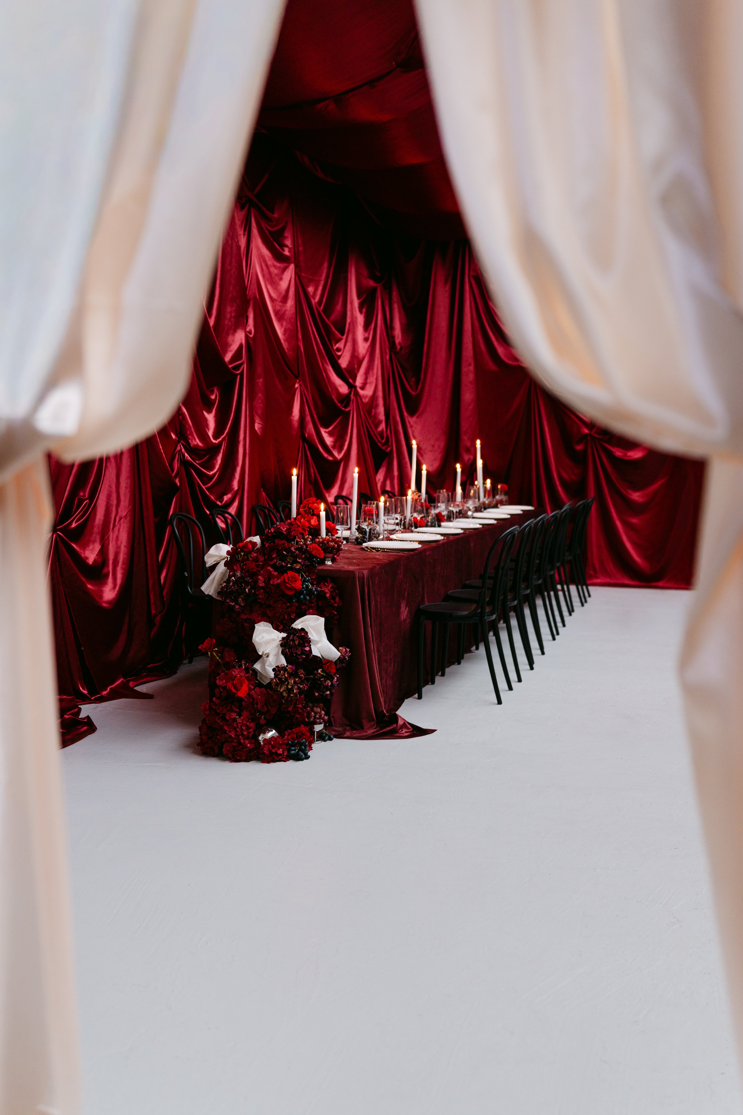 A long dining table decorated with red drapes, candles, and floral arrangements, viewed through a doorway with beige curtains.