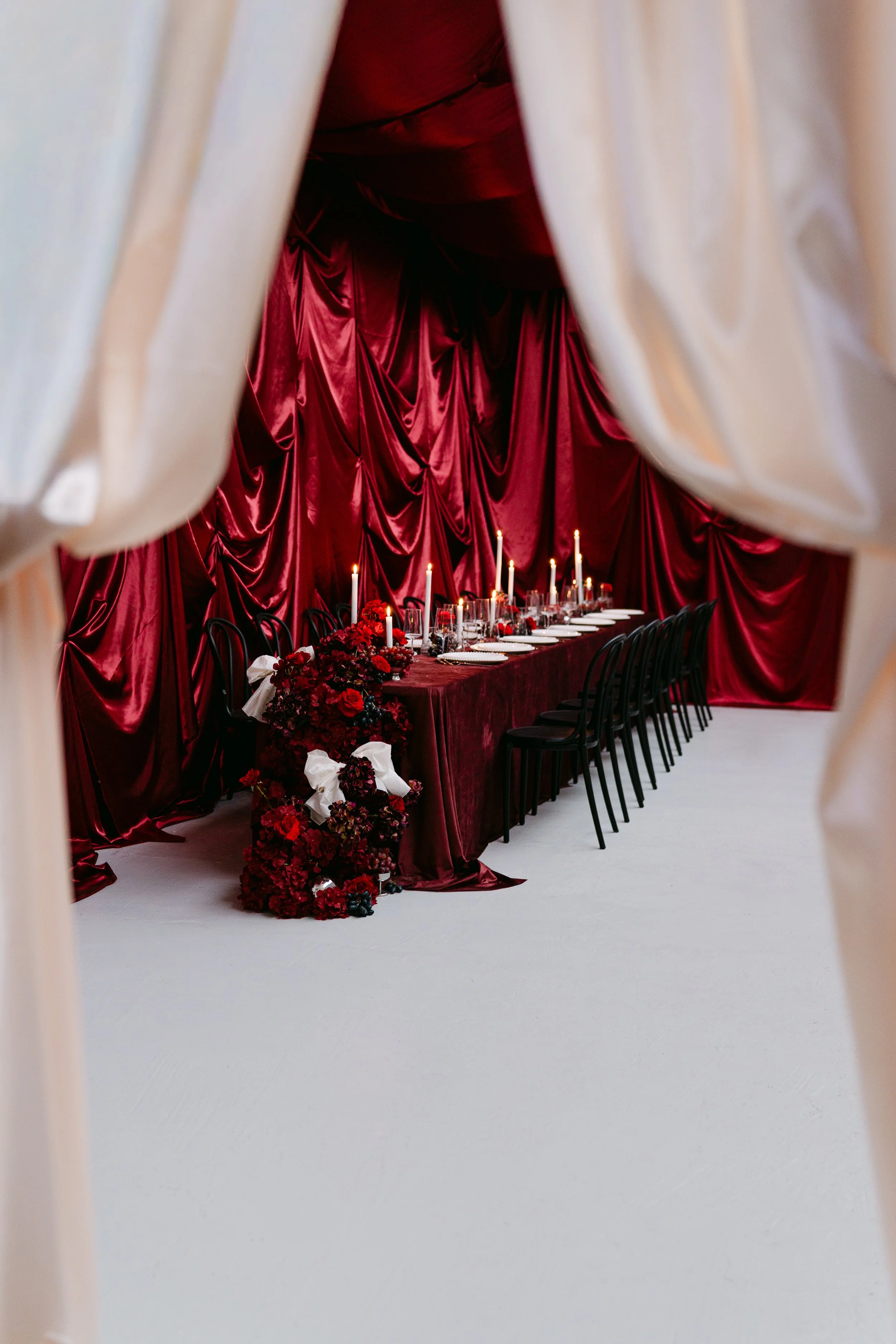 A long dining table decorated with red drapes, candles, and floral arrangements, viewed through a doorway with beige curtains.