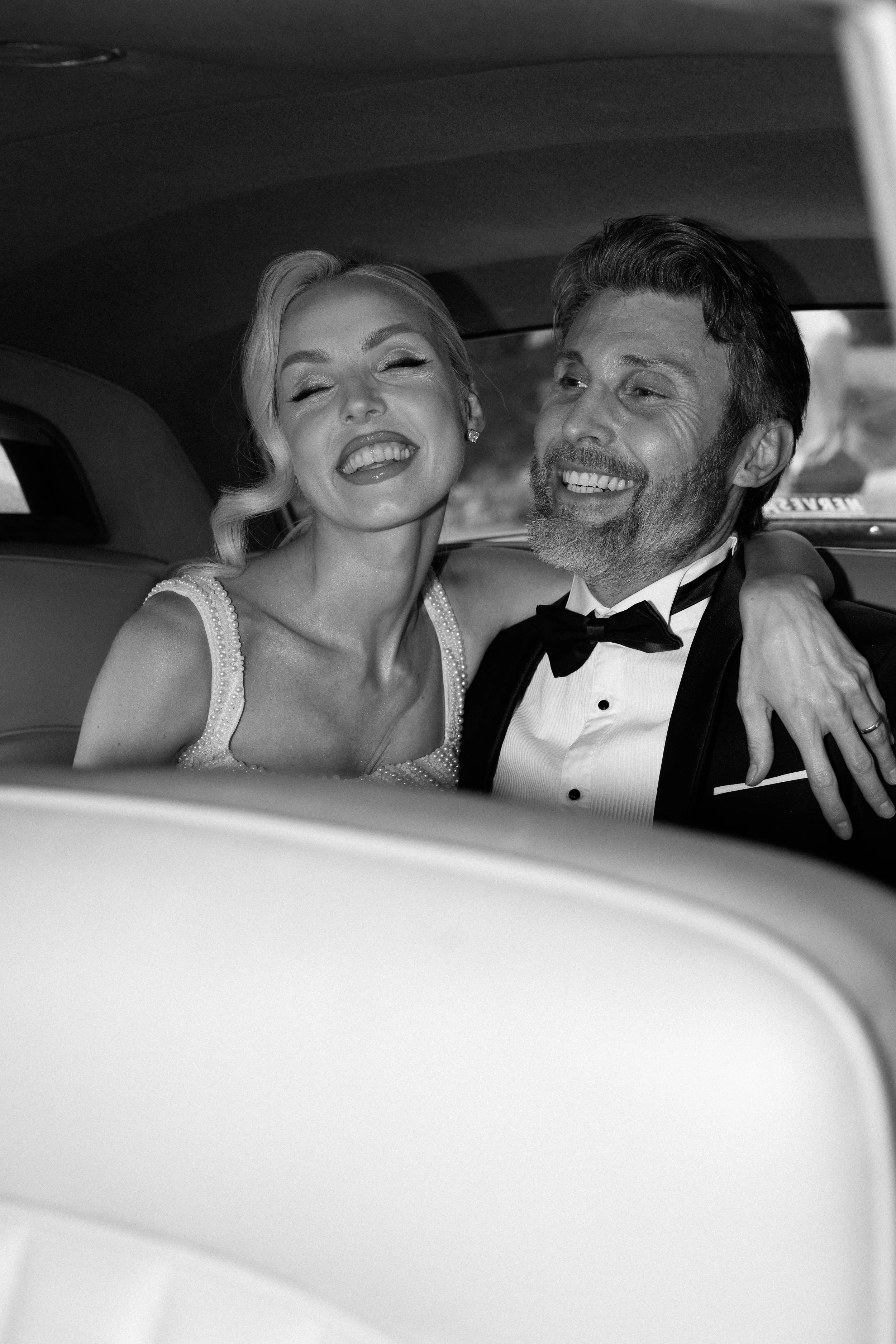 A black and white photo of a woman and man dressed in formal attire sitting inside a car, smiling and enjoying a joyful moment.