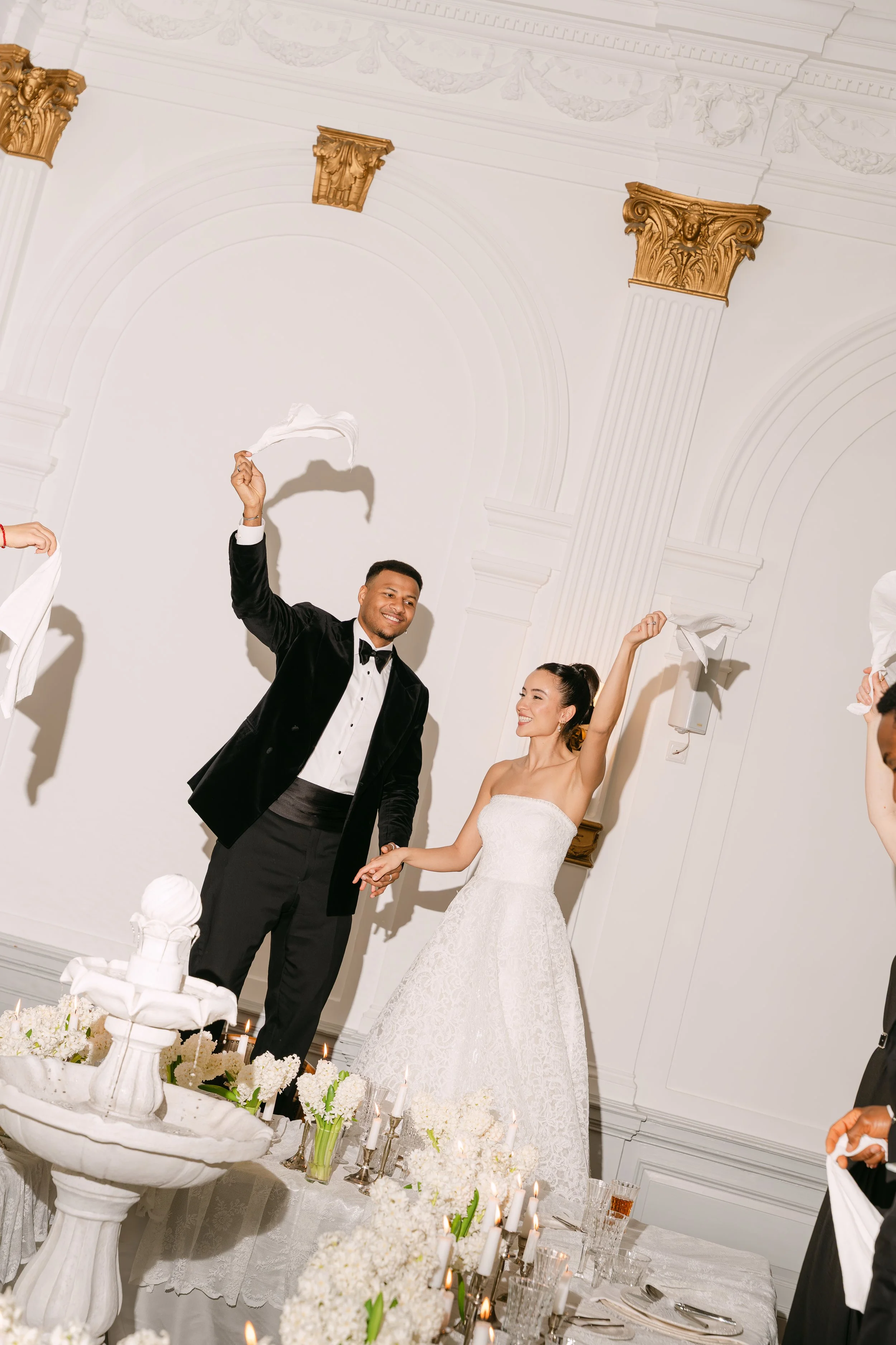 A newlywed couple at their wedding reception, holding hands and smiling. The groom is wearing a black tuxedo with a bow tie, and the bride is in a white lace wedding gown. They are dancing in a decorated elegant room with white walls, gold accents, a