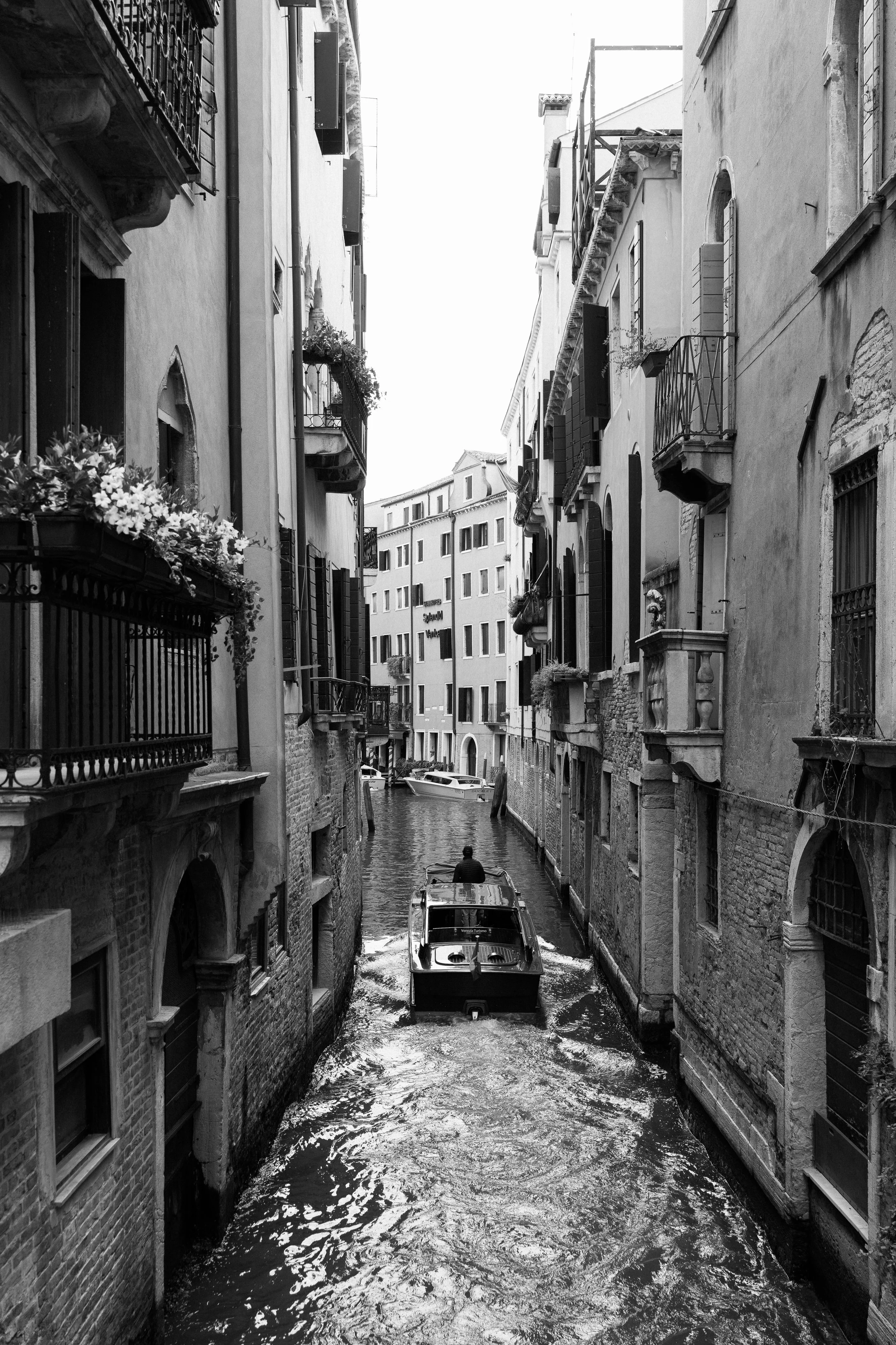 A black and white photo of a narrow canal in Venice, Italy, with a small motorboat carrying a person along the waterway, surrounded by tall old buildings with balconies and flower boxes.