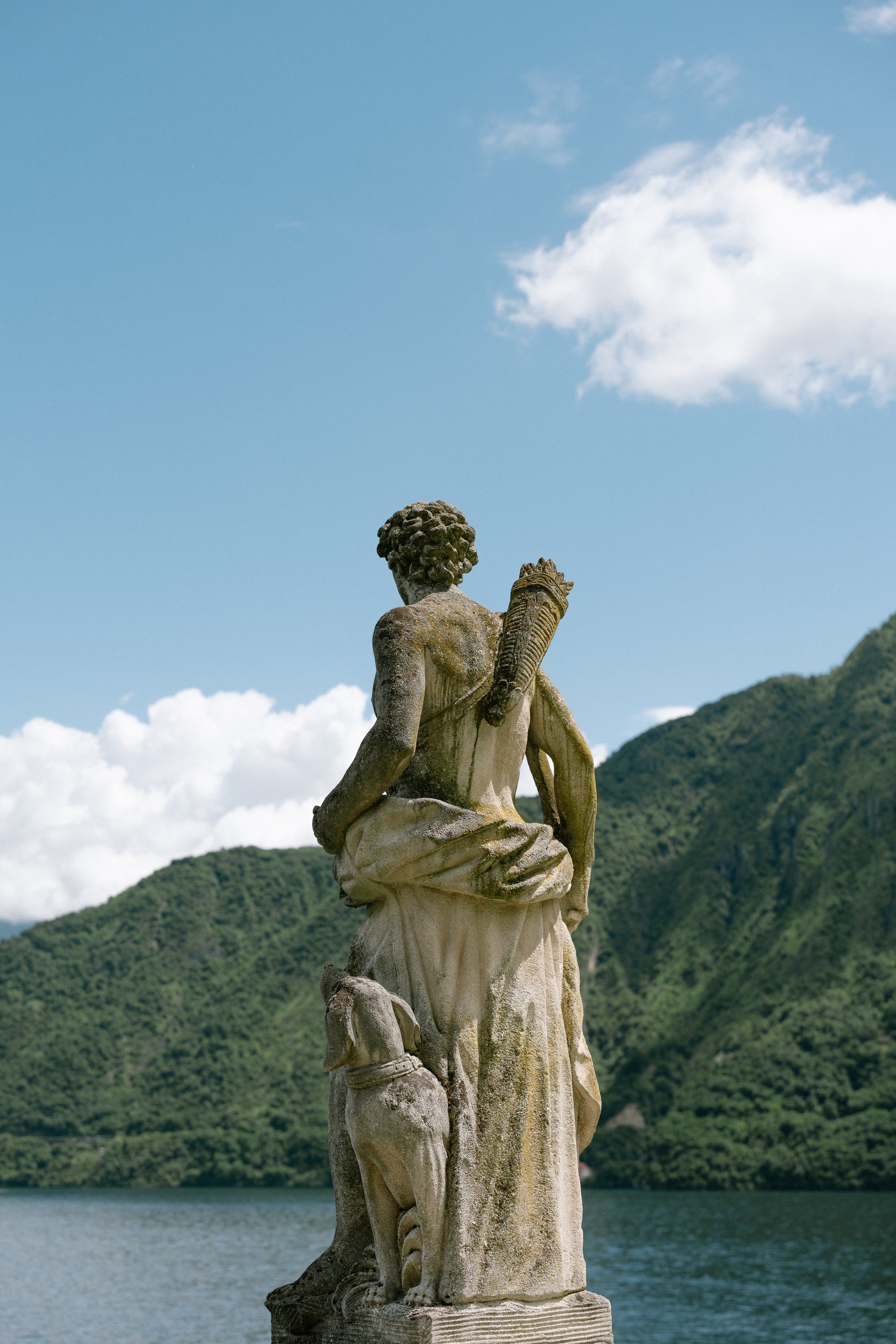 A weathered stone sculpture of a woman with a dog at her feet, set outdoors with mountains, a lake, and blue sky with clouds in the background.