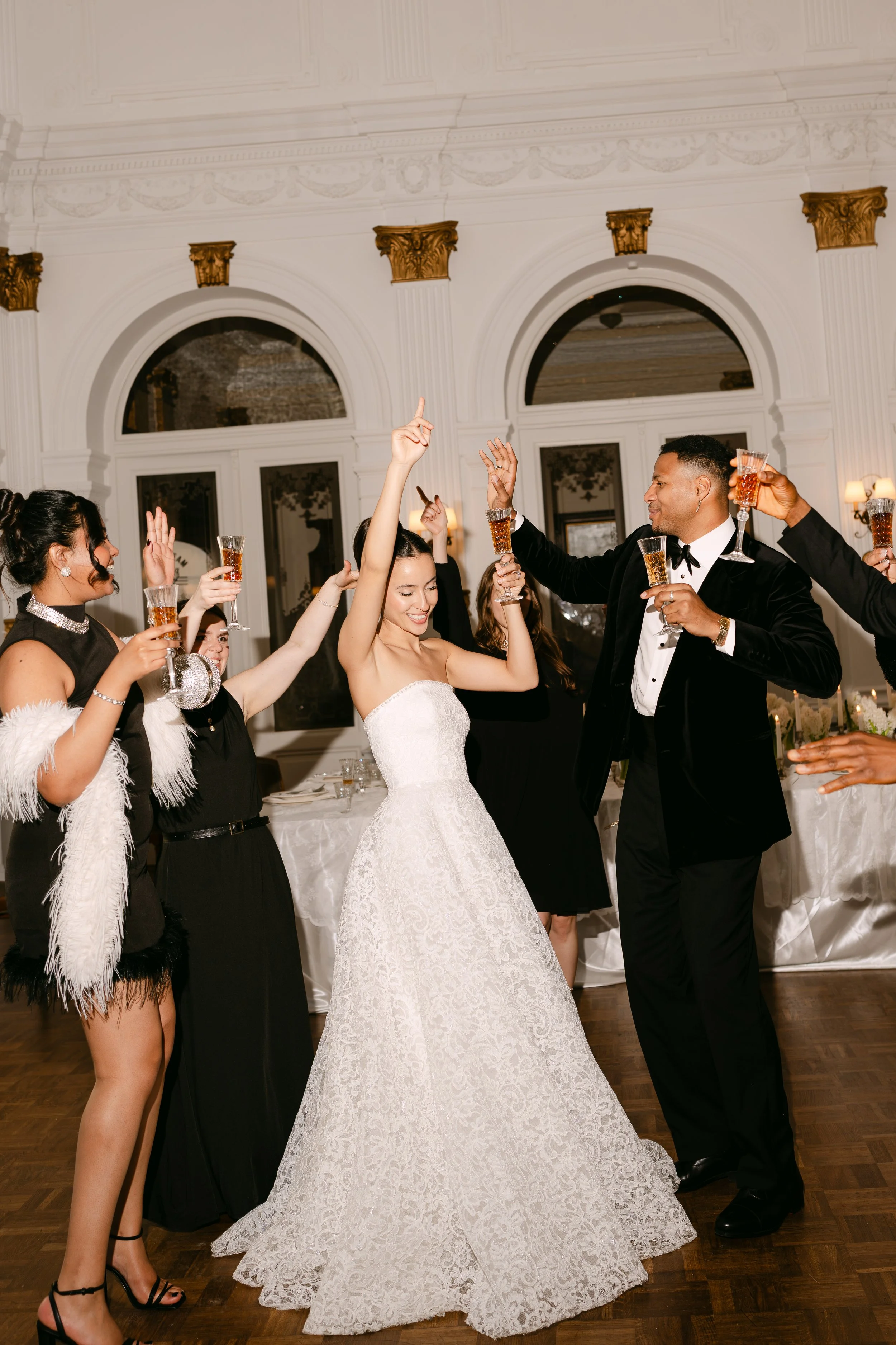 Couple dancing at a wedding reception with guests raising glasses for a toast, inside an elegant venue with white walls and gold accents.