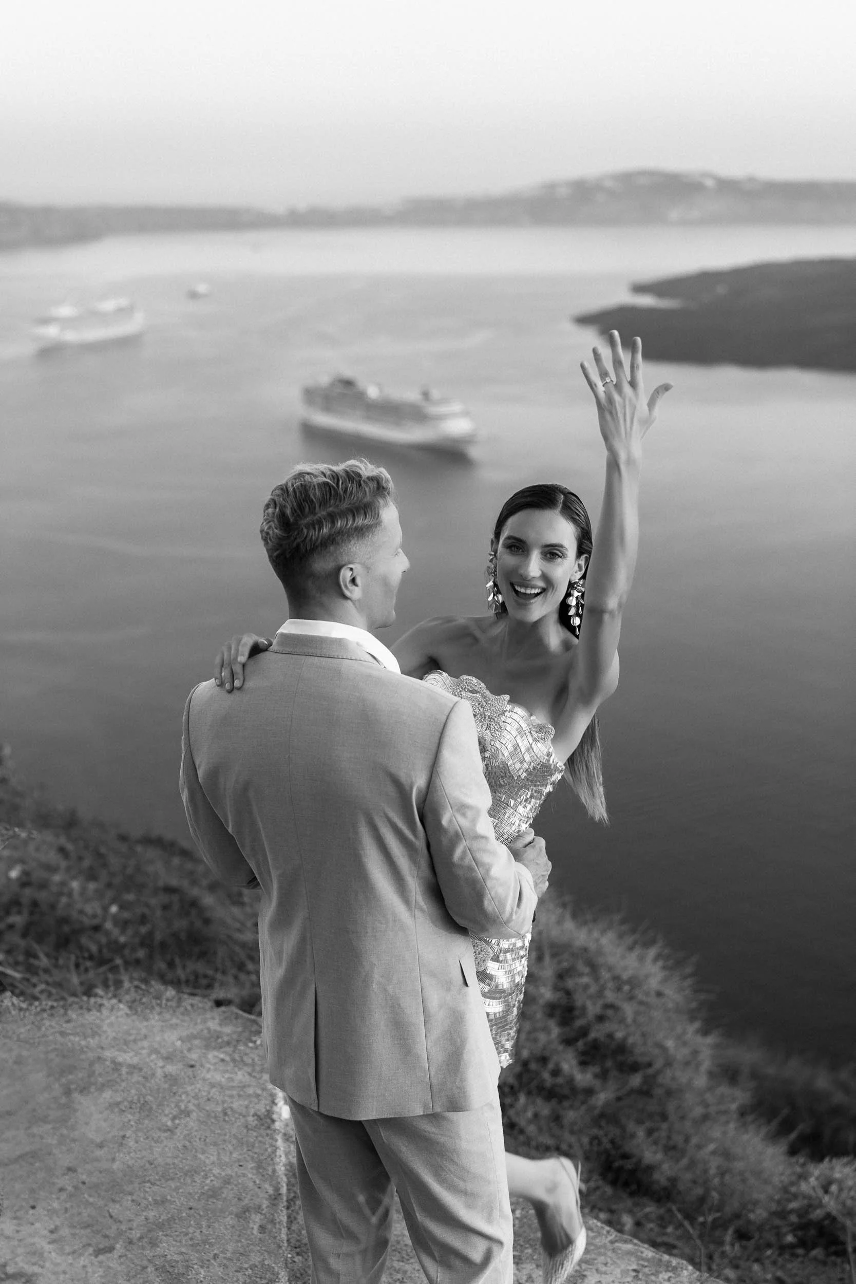 Black and white editorial wedding photography of a joyful bride showing her engagement ring while being held by her groom on a cliffside in Santorini, Greece. Overlooking the Aegean Sea and caldera, capturing a luxury destination wedding vibe.