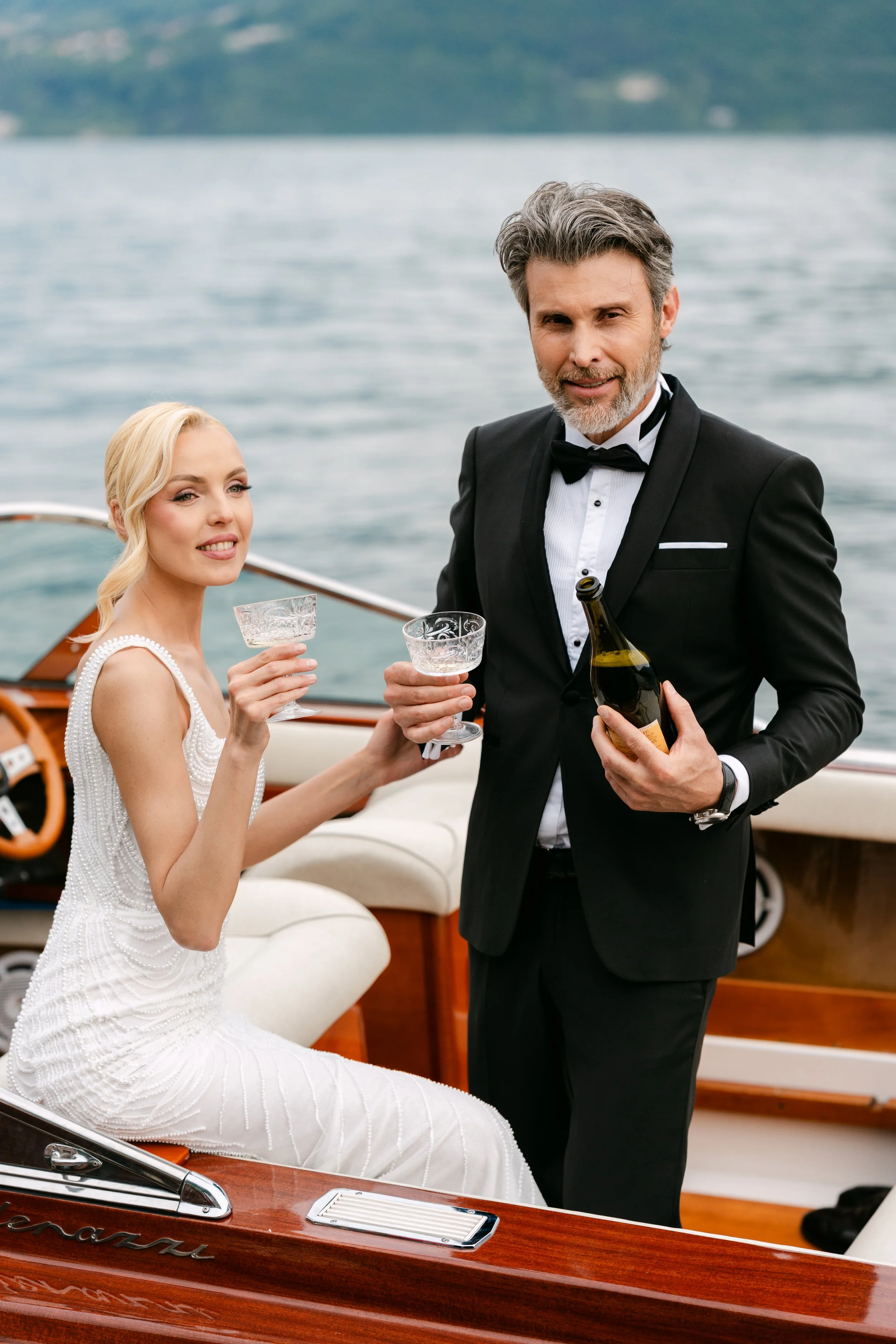 A man and woman dressed in formal attire on a boat, holding glasses and a bottle of champagne, with water in the background.