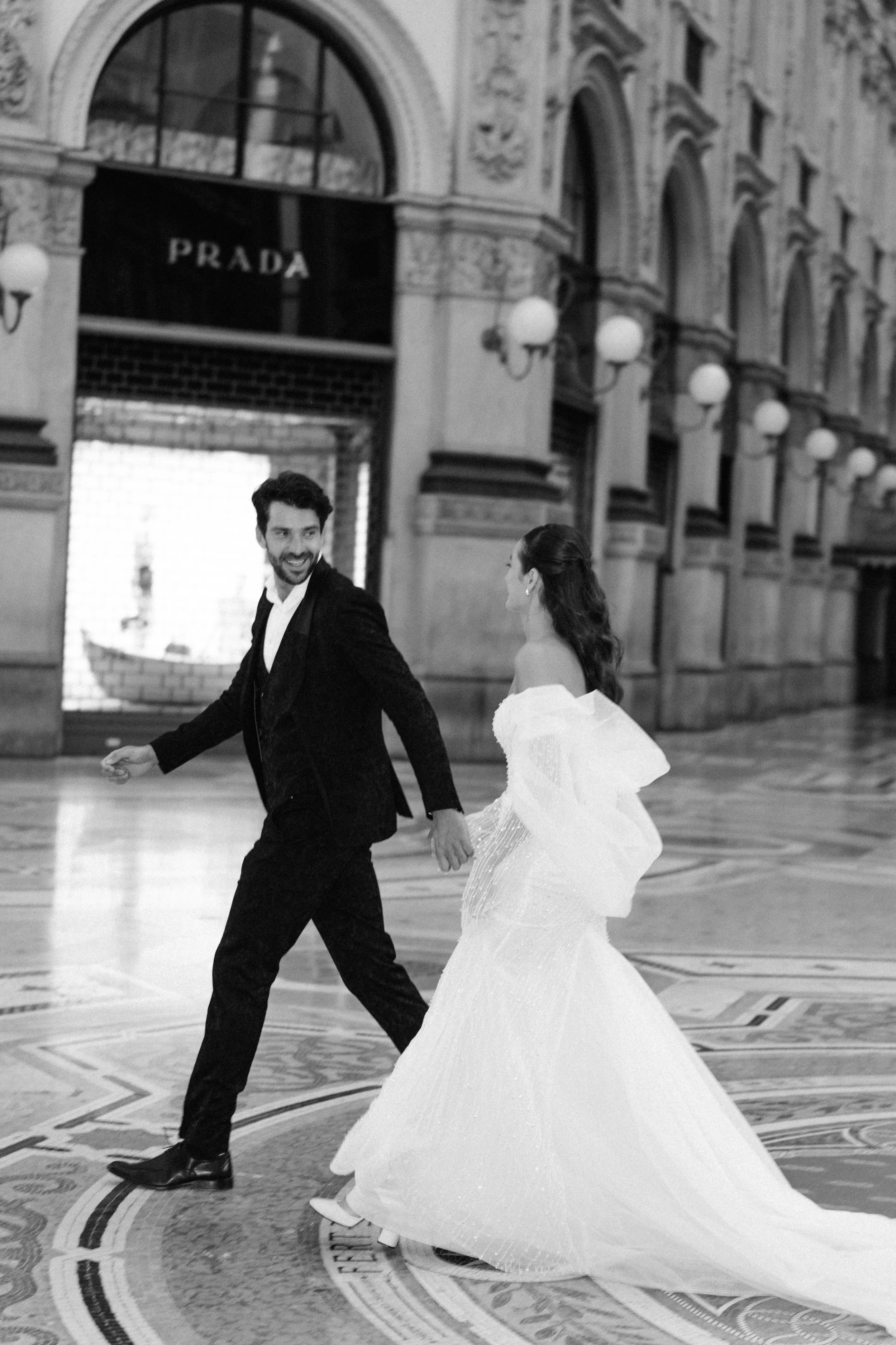A black and white photo of a smiling couple in wedding attire holding hands and dancing in an elegant, ornate indoor setting with high arched windows and historic architectural details.
