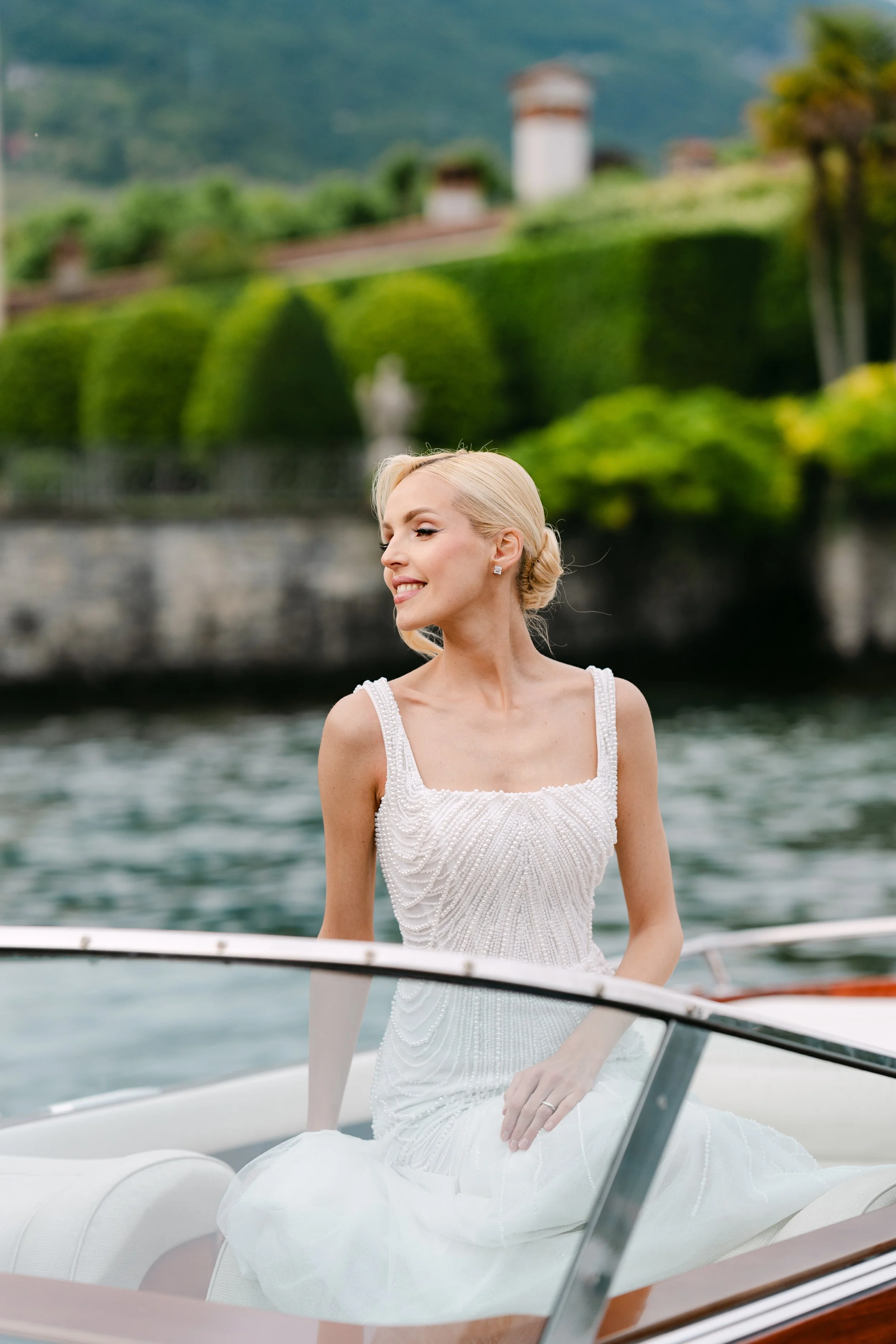 A woman in a wedding dress sitting on a boat with a scenic hillside and greenery in the background.