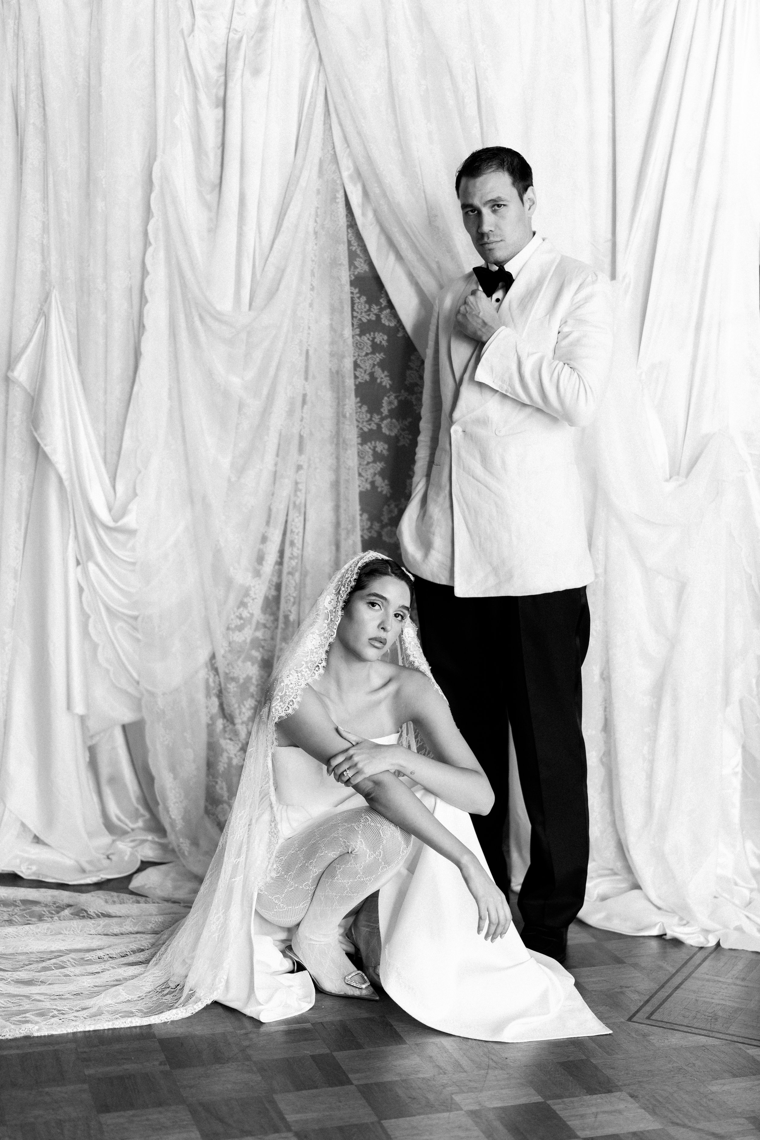 A black-and-white photograph of a man in a tuxedo and a woman in a bridal gown with a veil. The man is standing while adjusting his tuxedo, and the woman is sitting on the floor, looking at the camera, with draped curtains in the background.