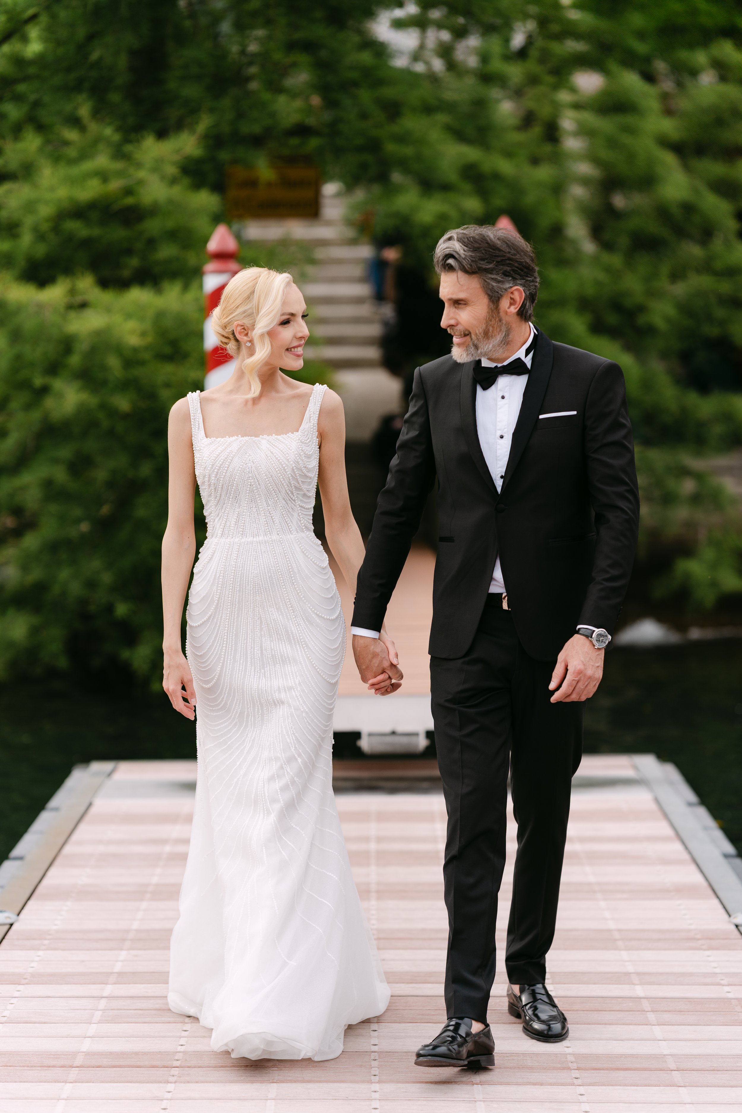 A bride and groom holding hands and walking on a wooden dock by a body of water, surrounded by green trees, during their wedding.