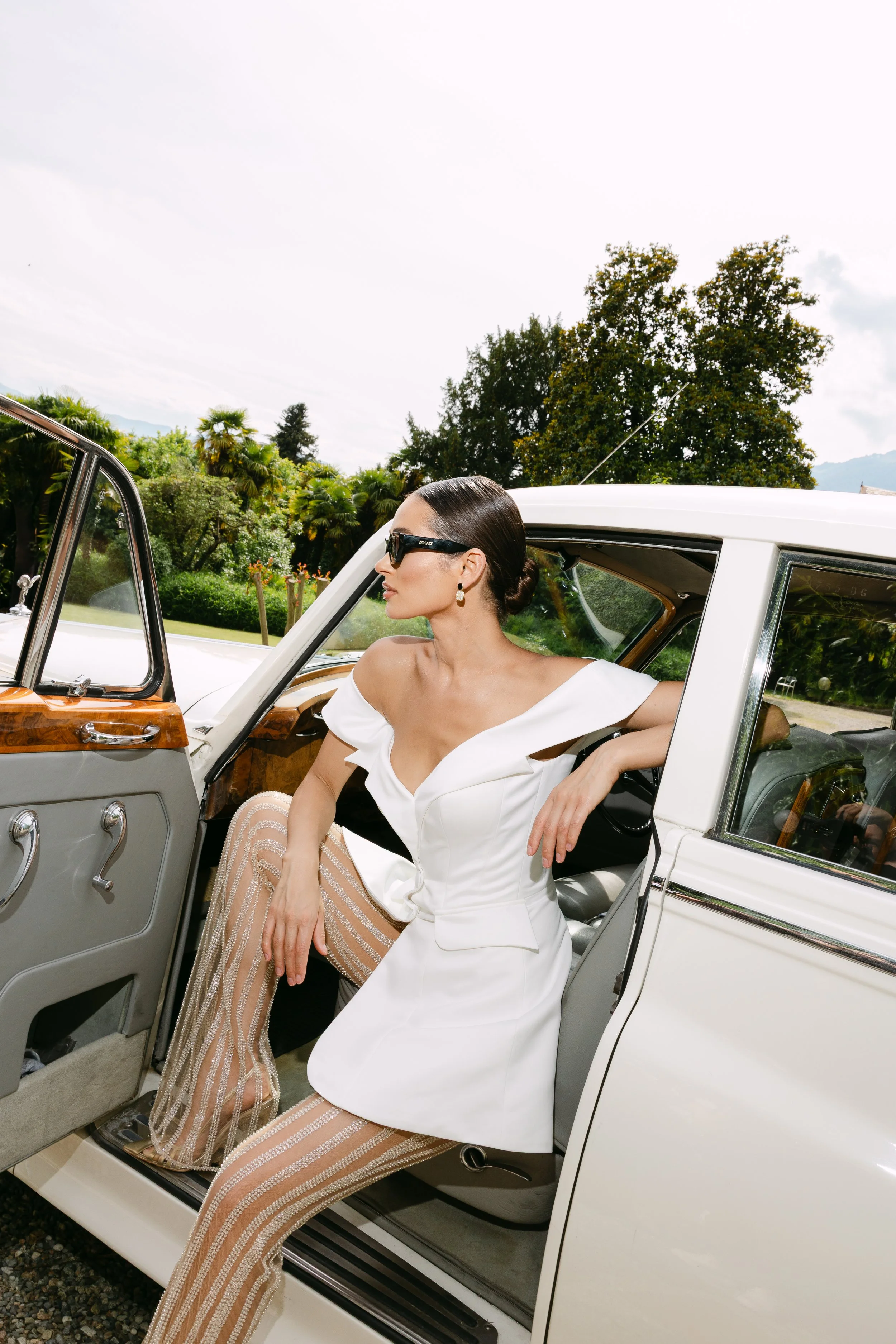 Woman in white dress and sunglasses sitting in an open vintage car with a scenic green landscape and trees in the background.