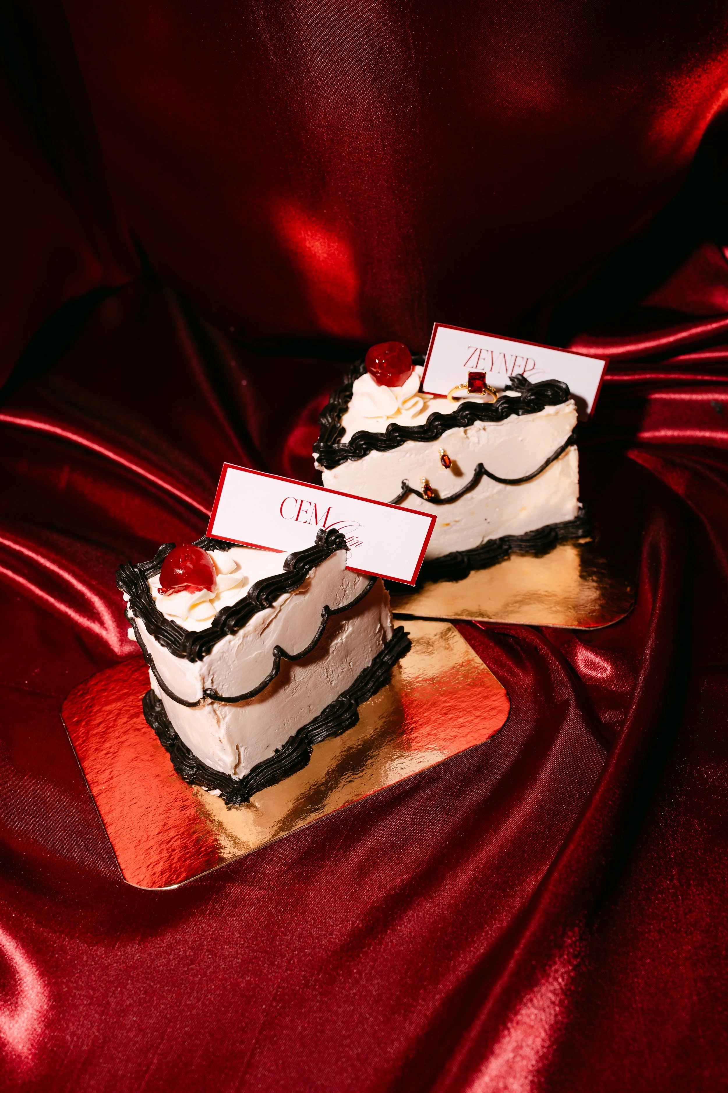Two slices of cake decorated with black and white frosting, cherries, and small gold accents, placed on gold-colored bases on red satin fabric.