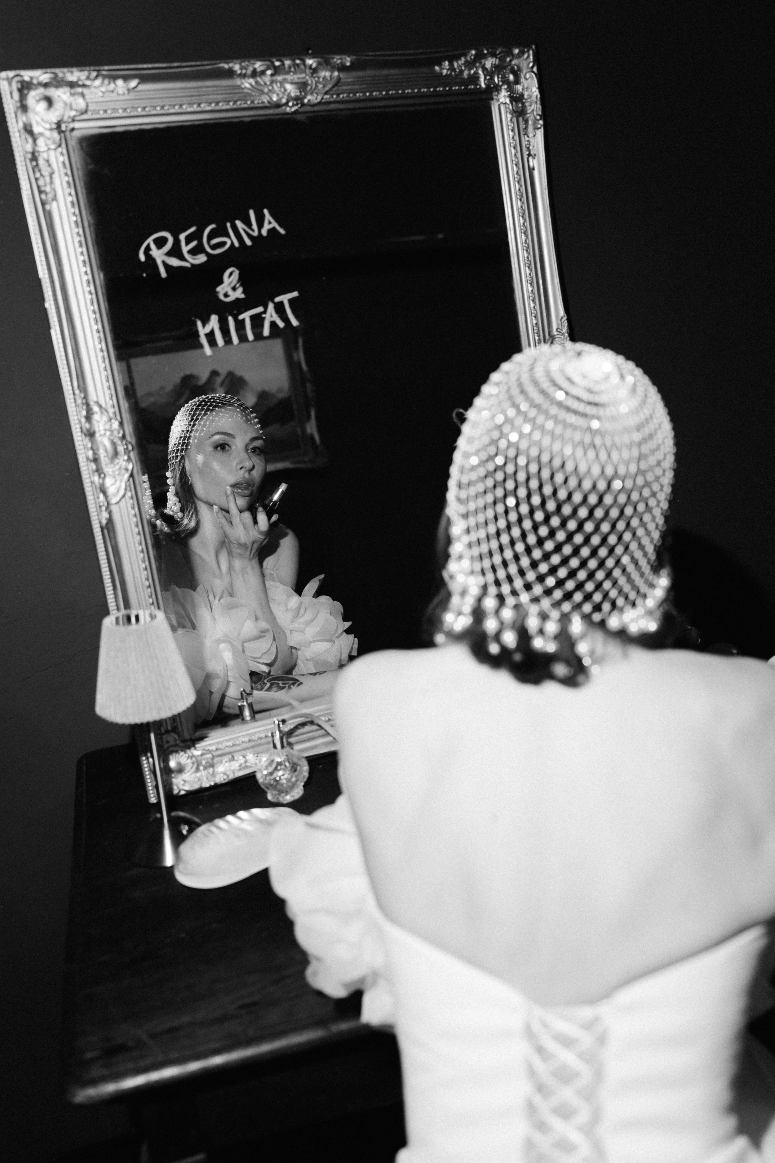 A woman wearing a pearl headband and earrings looking into a mirror. She is applying makeup or lipstick, with her finger near her lips. The mirror has a decorative ornate frame and a sign that reads 'Regina & Mitat'. The scene is in a dimly lit room 
