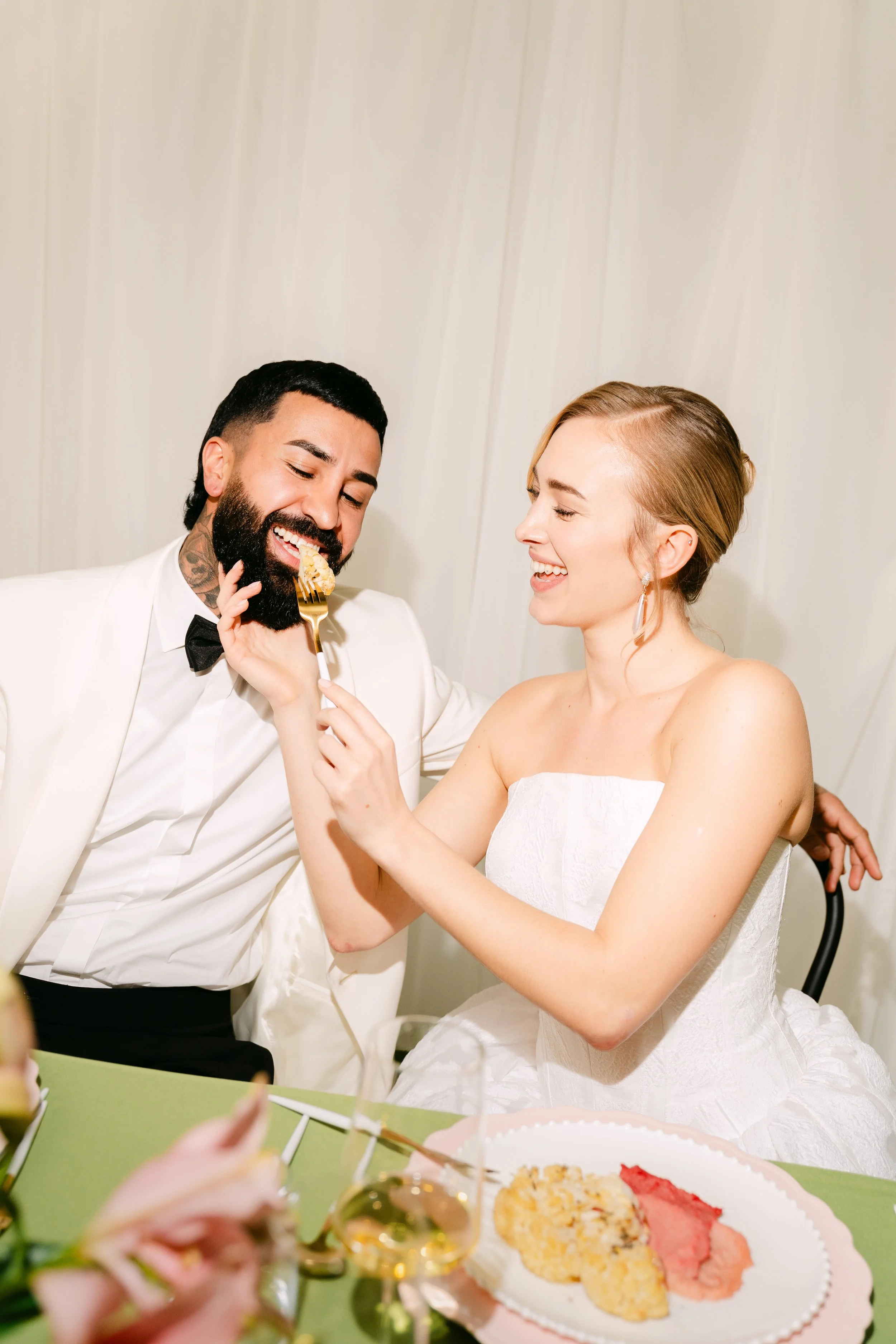 A man in a white tuxedo and a woman in a white dress are smiling as the man eats a slice of cake given to him by the woman. They are at a celebration, with a table of food and decorations visible in the foreground.