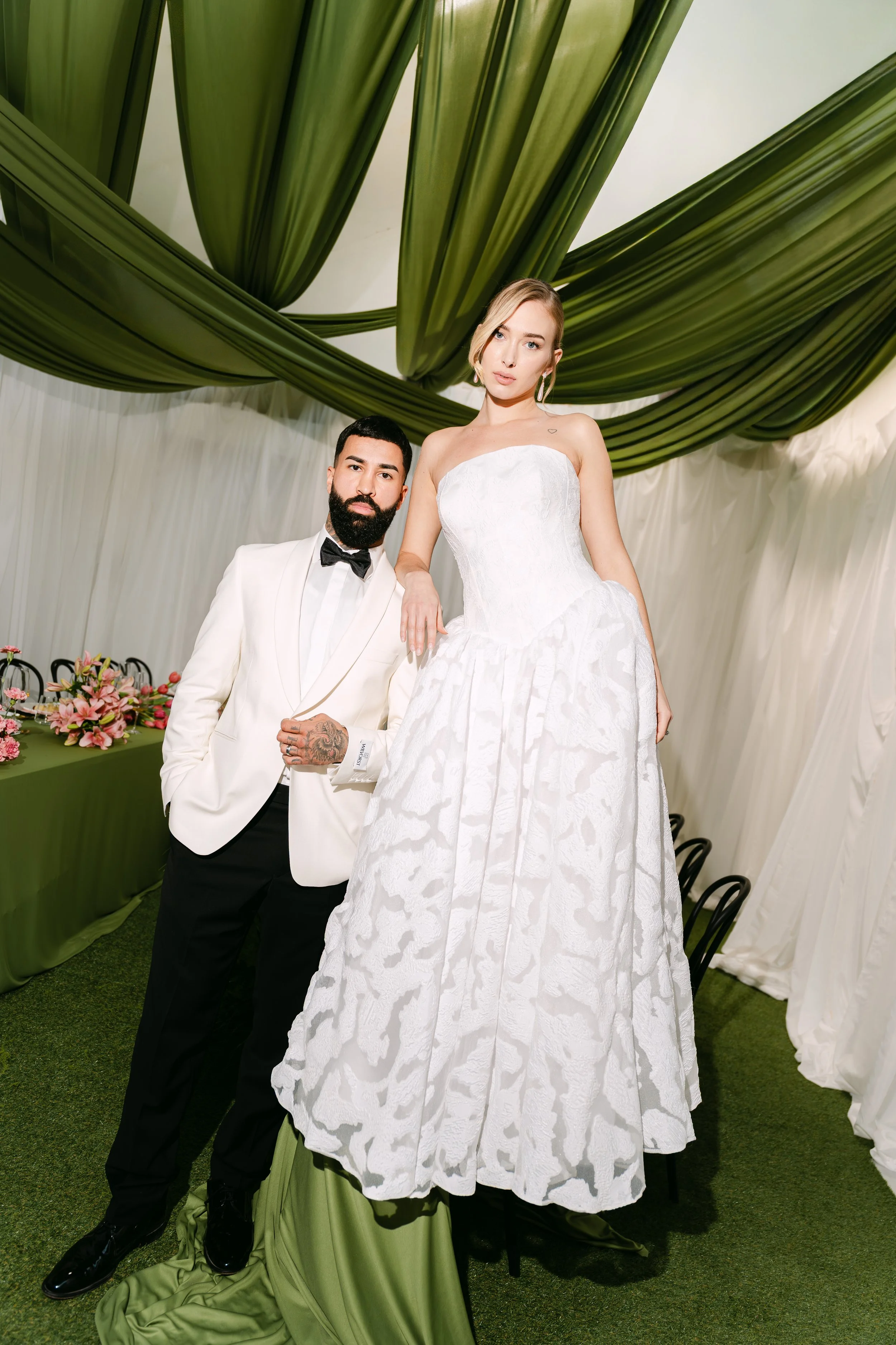 A man and woman in wedding attire posing at a wedding reception. The woman is wearing a strapless white wedding gown, and the man is in a white tuxedo jacket with black pants and a black bowtie. They are standing under a decorative ceiling with green