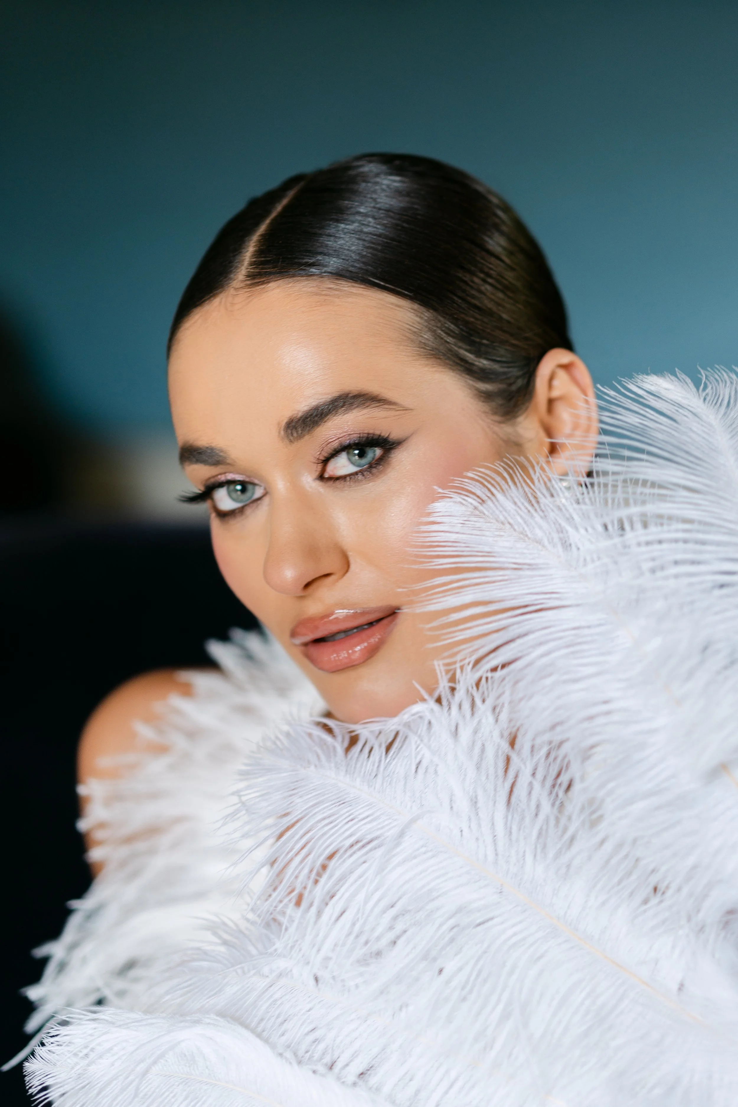 A woman with dark hair styled in a sleek bun, blue eyes, and makeup, is holding white feathers near her face against a blue background.