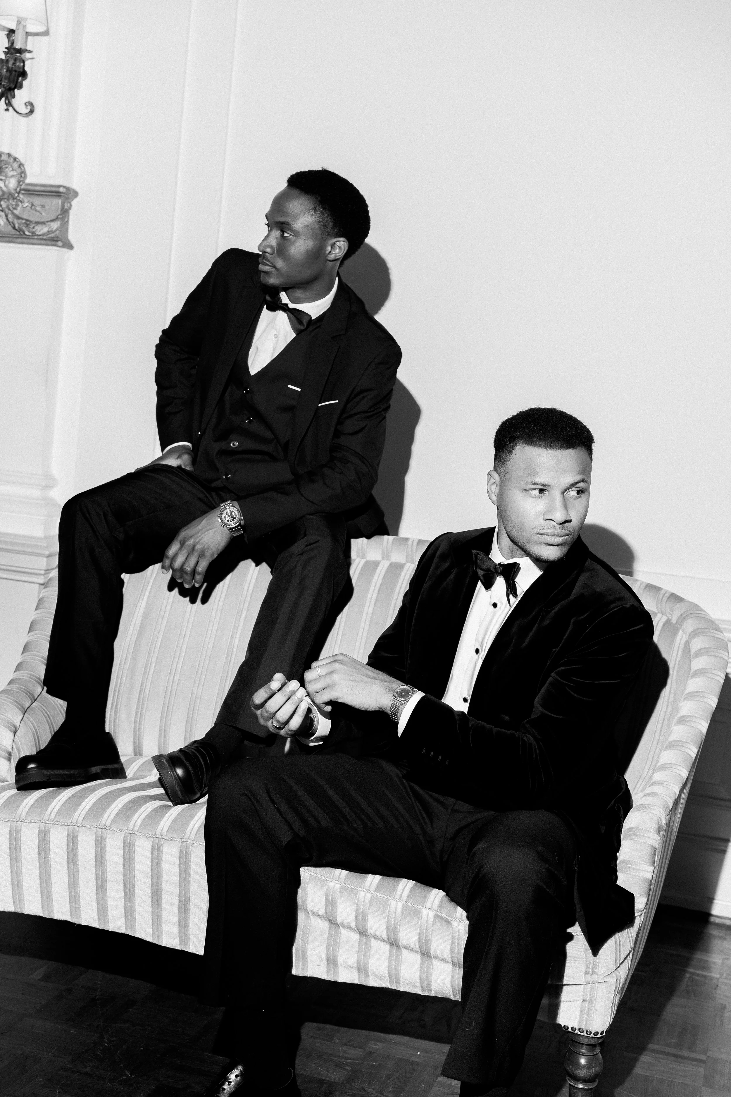 Two men dressed in formal tuxedos sitting on a striped sofa in a room with elegant decor. The man on the left is sitting with one leg crossed over the other, wearing a watch and looking to his left. The man on the right is seated with his hands clasp