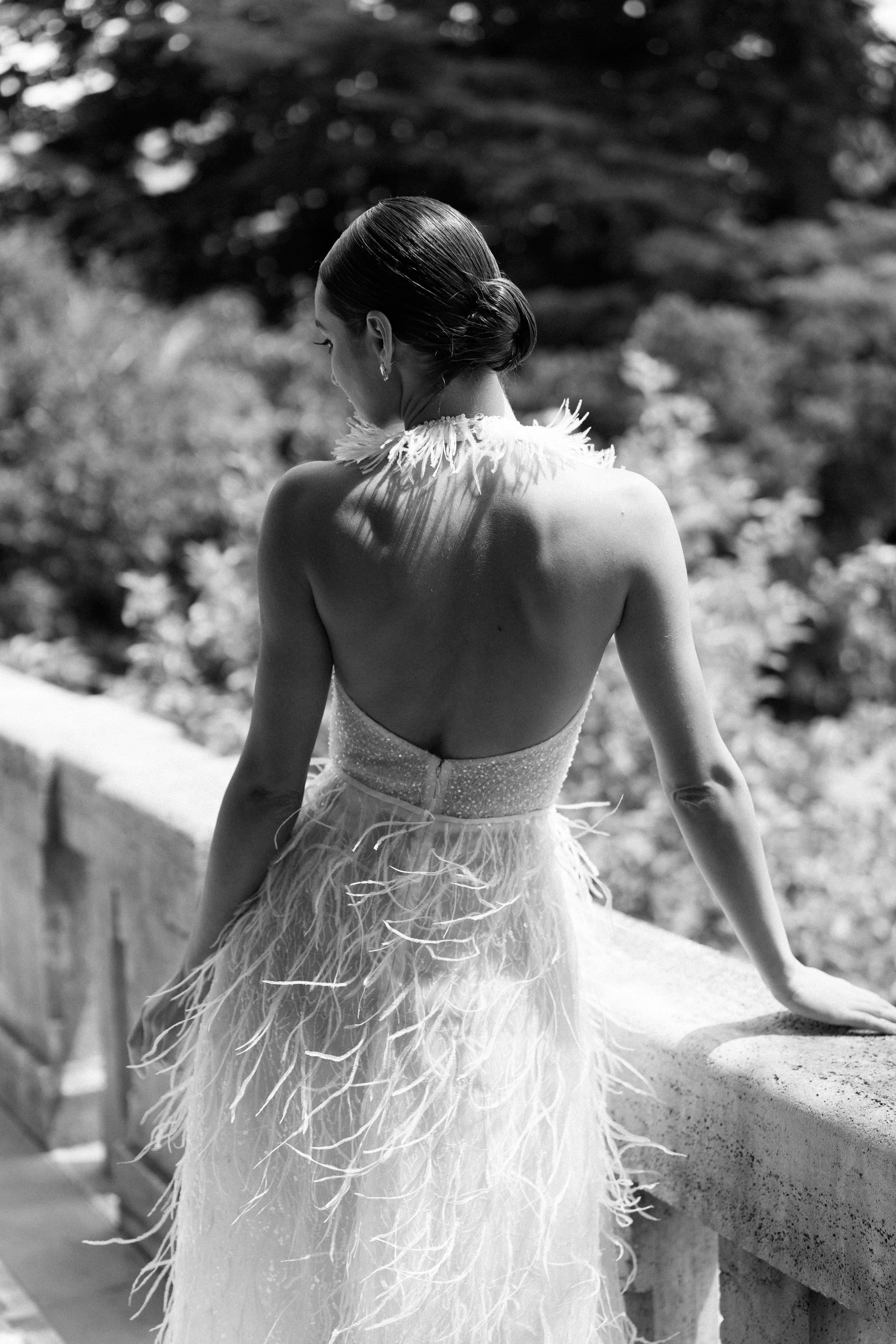 Black and white photograph of a woman wearing an elegant, backless dress with feathered details, standing outdoors on a balcony with a blurred natural background.