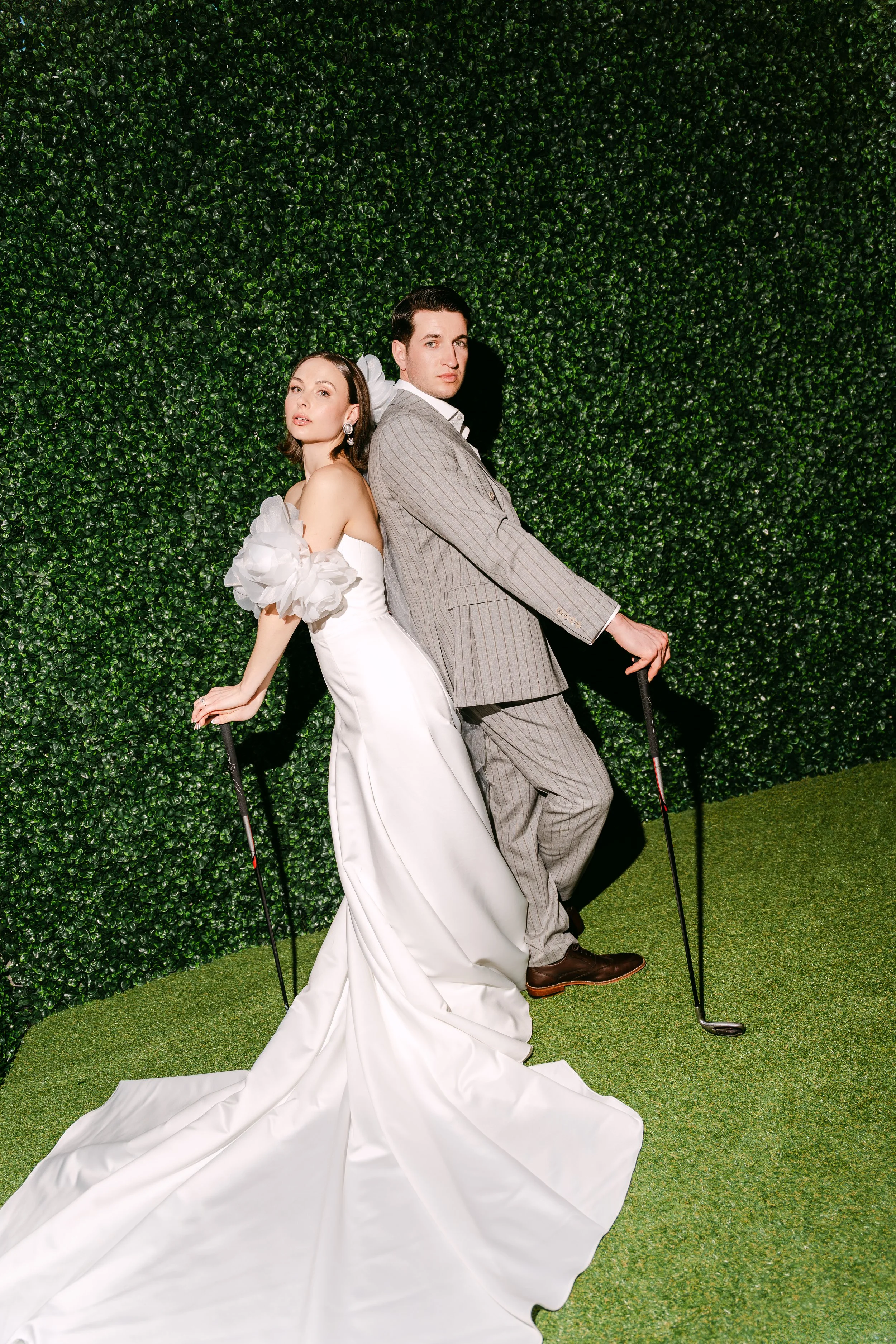 A man and woman dressed as golfers with golf clubs, standing back to back against a green leafy wall, on a grassy surface.