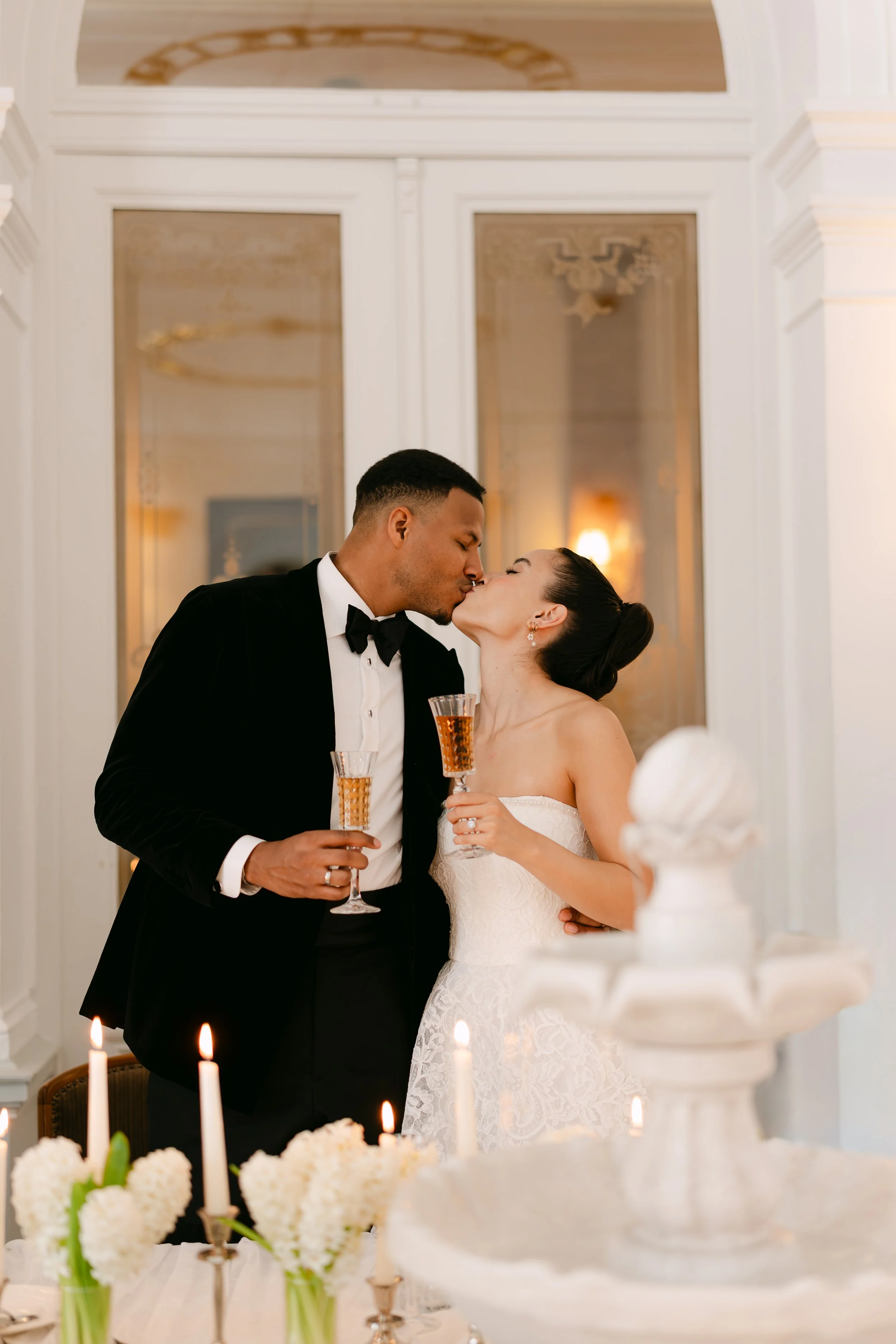 A bride and groom share a kiss at their wedding reception, holding champagne flutes, with wedding cake and candles on a decorated table in the foreground.