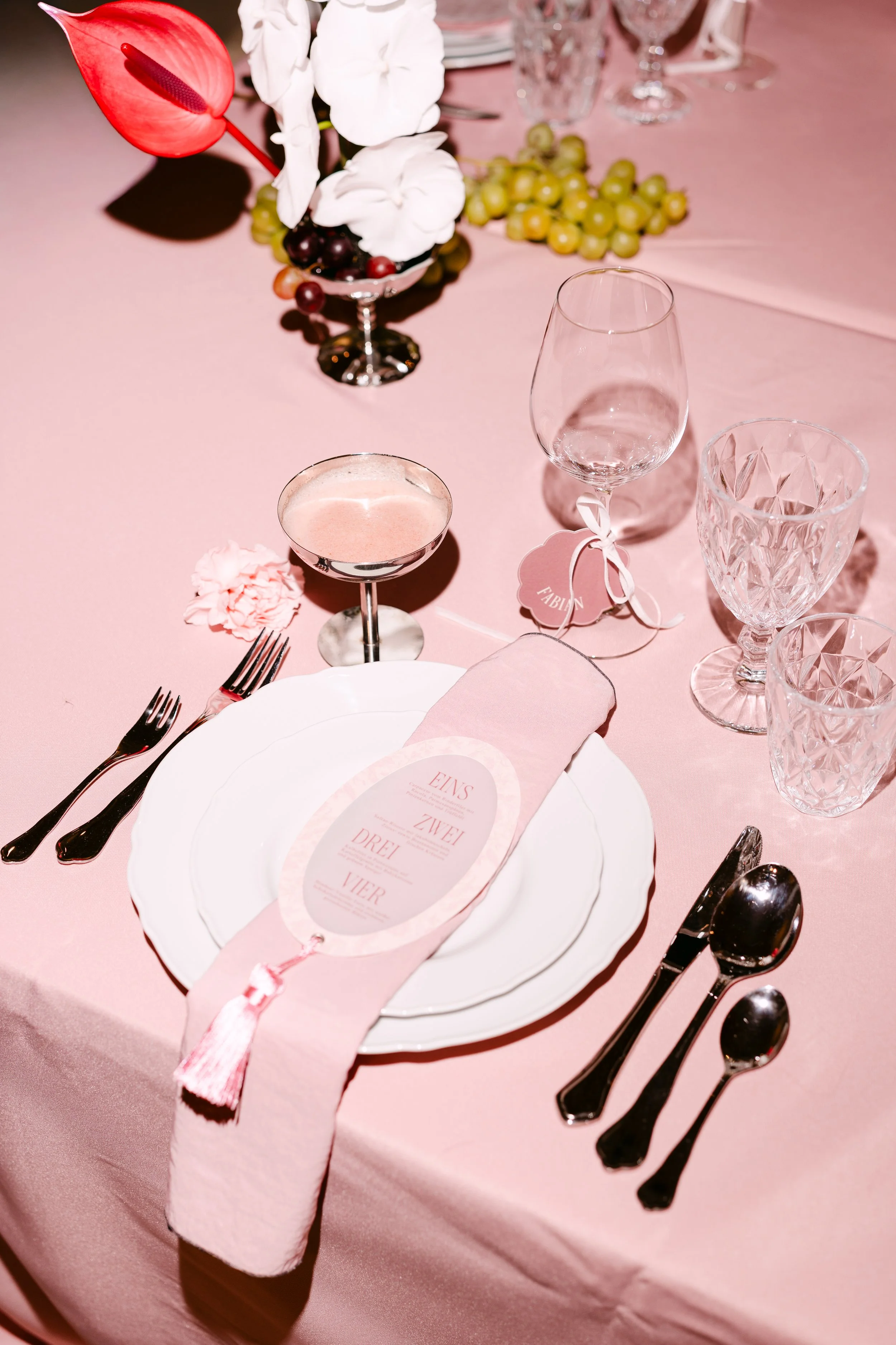 A pink table setting at a celebration with white plates, silverware, wine glasses, a pink and white menu, and a floral centerpiece with white flowers, anthurium, and grapes.