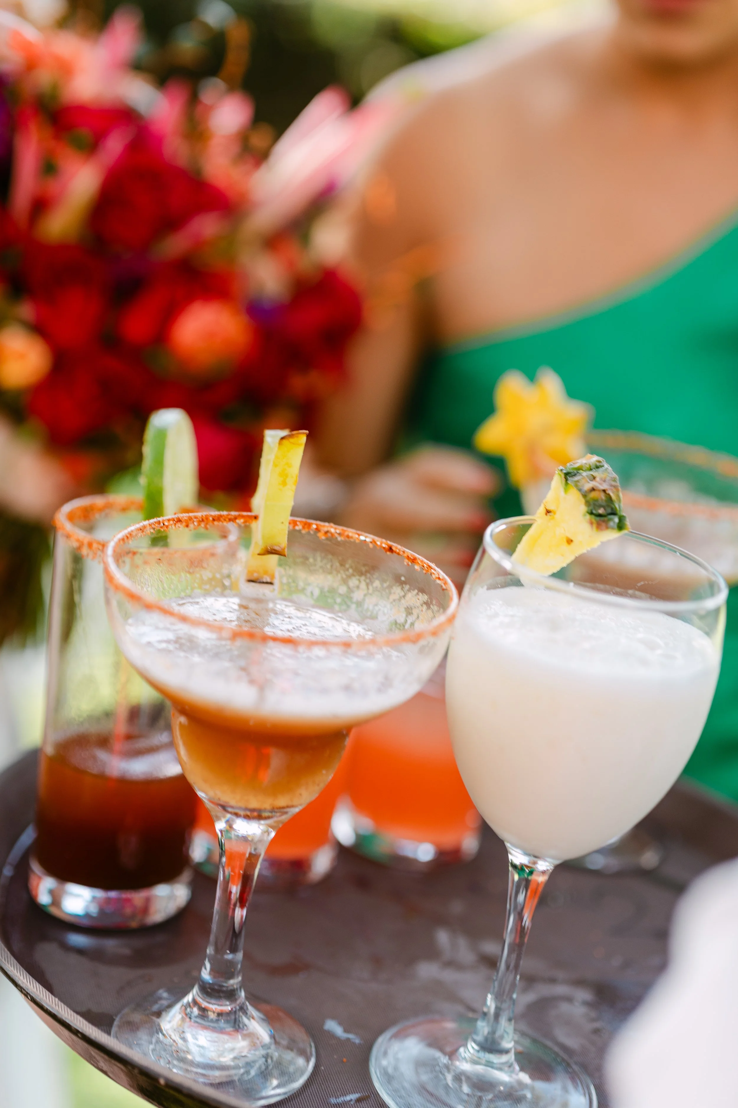 Several colorful cocktails on a tray, garnished with fruits like pineapple, lime, and celery stalks, with a blurred person and bouquet of flowers in the background.