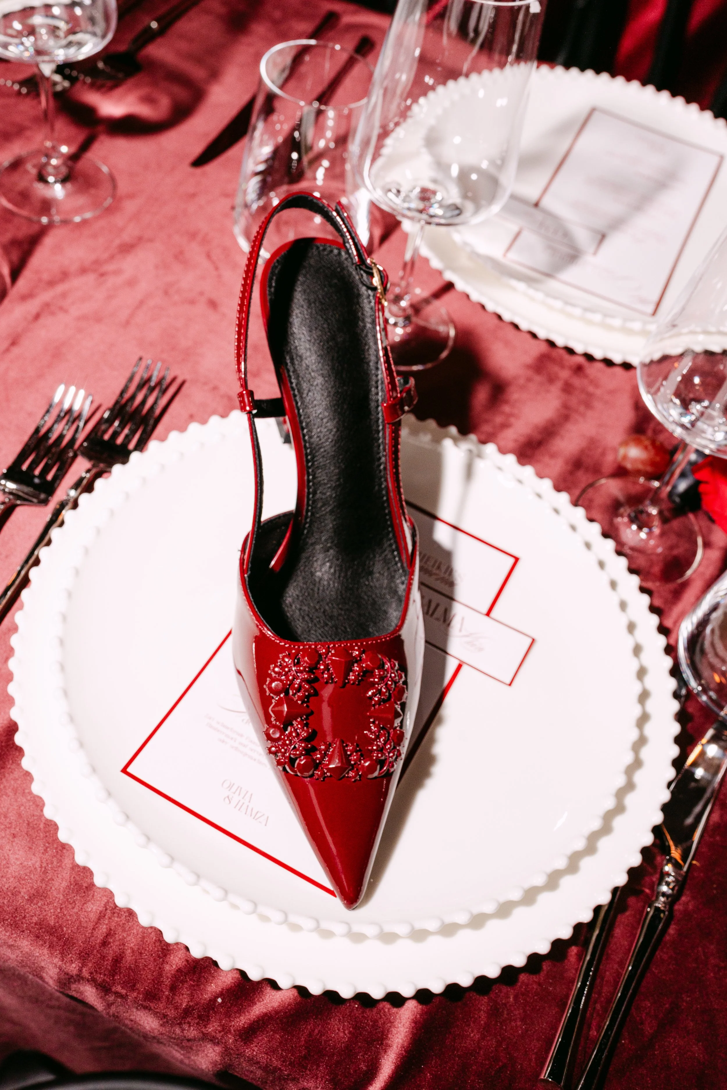 A red high-heeled shoe with decorative embellishments displayed on a white plate at a formal dining table with glassware, cutlery, and a burgundy tablecloth.