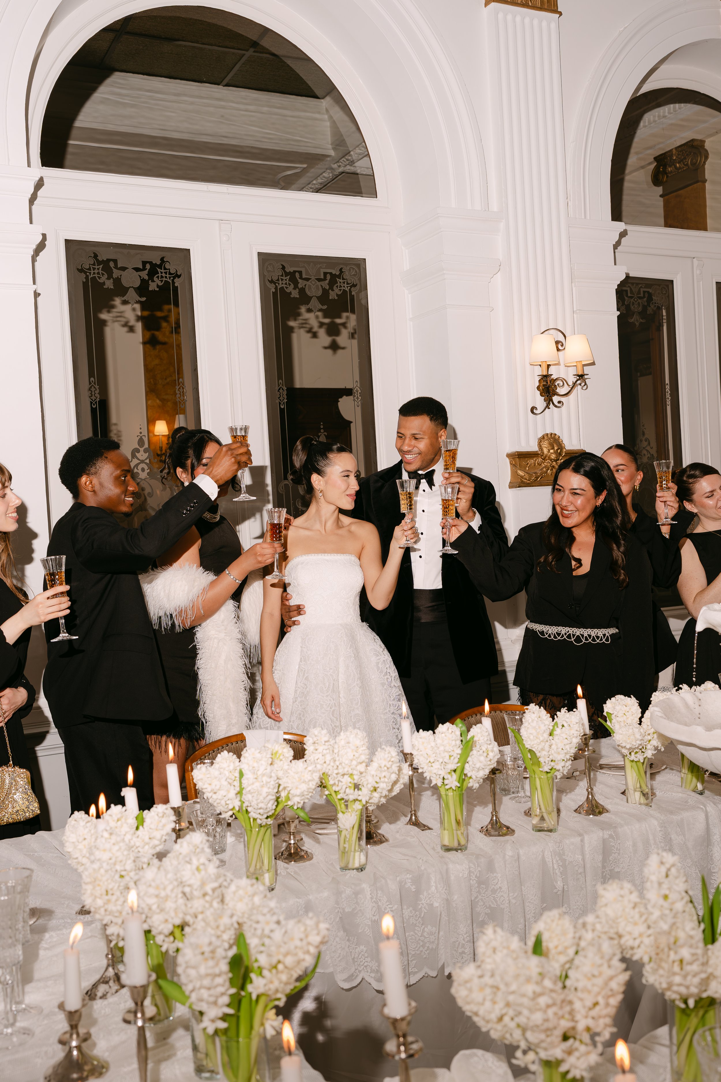 A wedding reception with a bride in a white wedding dress and groom in a tuxedo, surrounded by guests raising glasses in a toast, in a decorated hall with floral centerpieces and candles on the table.