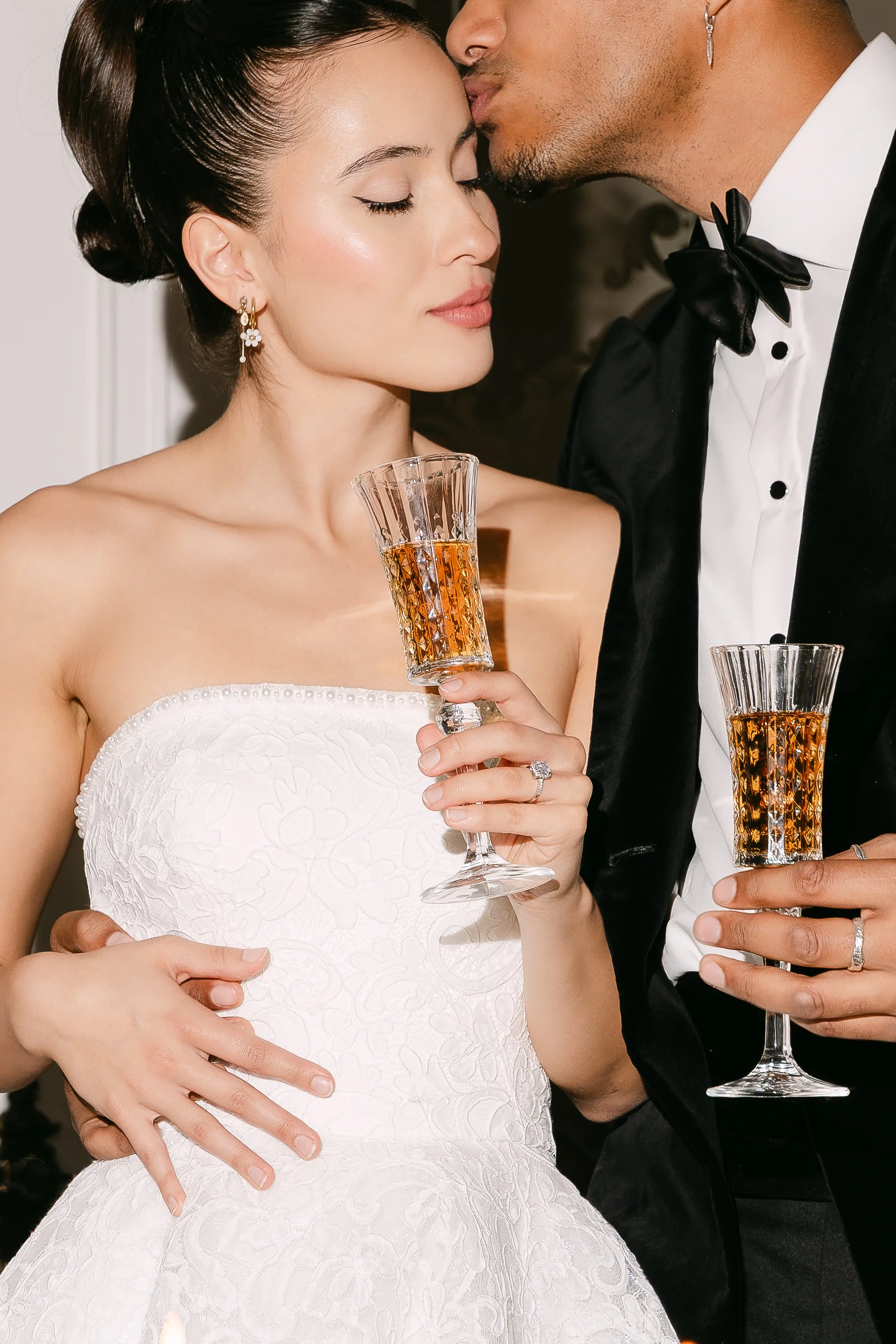 A bride and groom at their wedding, sharing a kiss on the forehead while holding glasses of champagne. The bride is in a white wedding dress with floral embroidery and gold earrings, and the groom is in a tuxedo with a black bow tie.