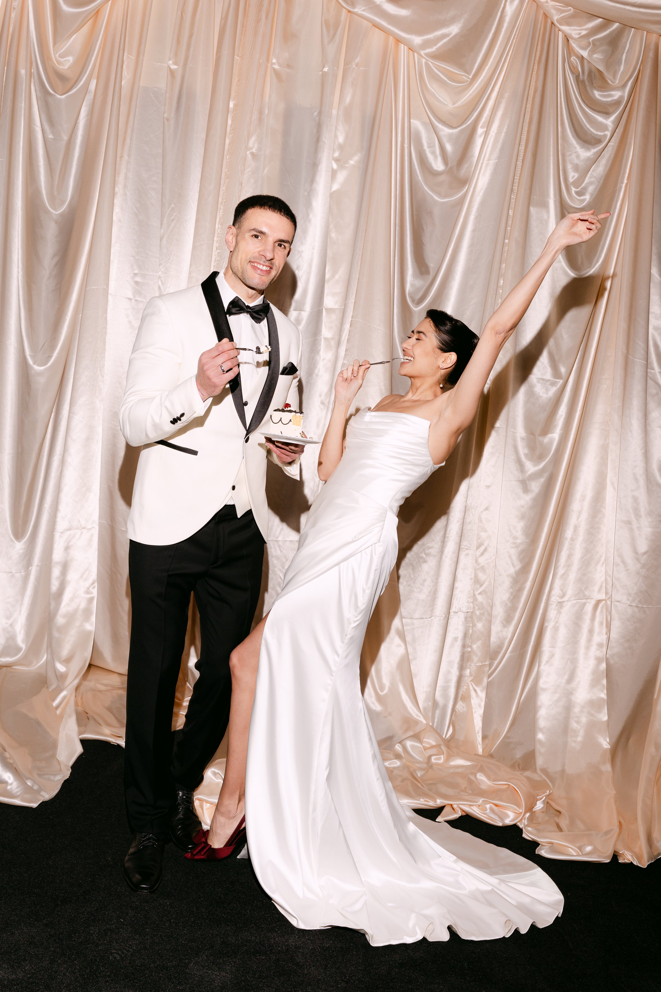 A bride and groom are celebrating at their wedding reception, dressed in formal attire, with the bride in a white gown and the groom in a tuxedo, standing in front of a satin curtain backdrop.