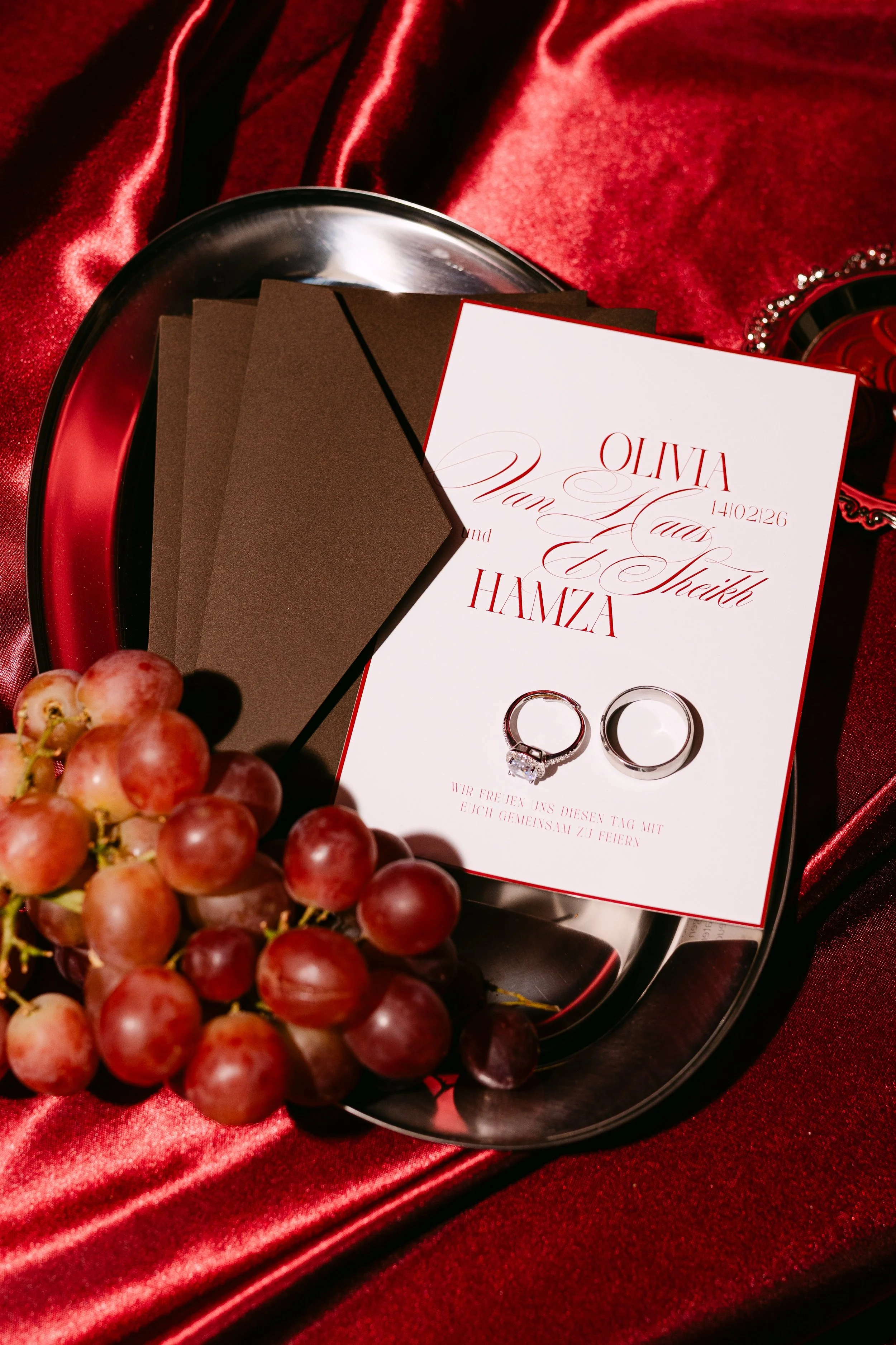 Wedding invitation card with two rings, a bunch of red grapes, and a silver tray on red velvet fabric.