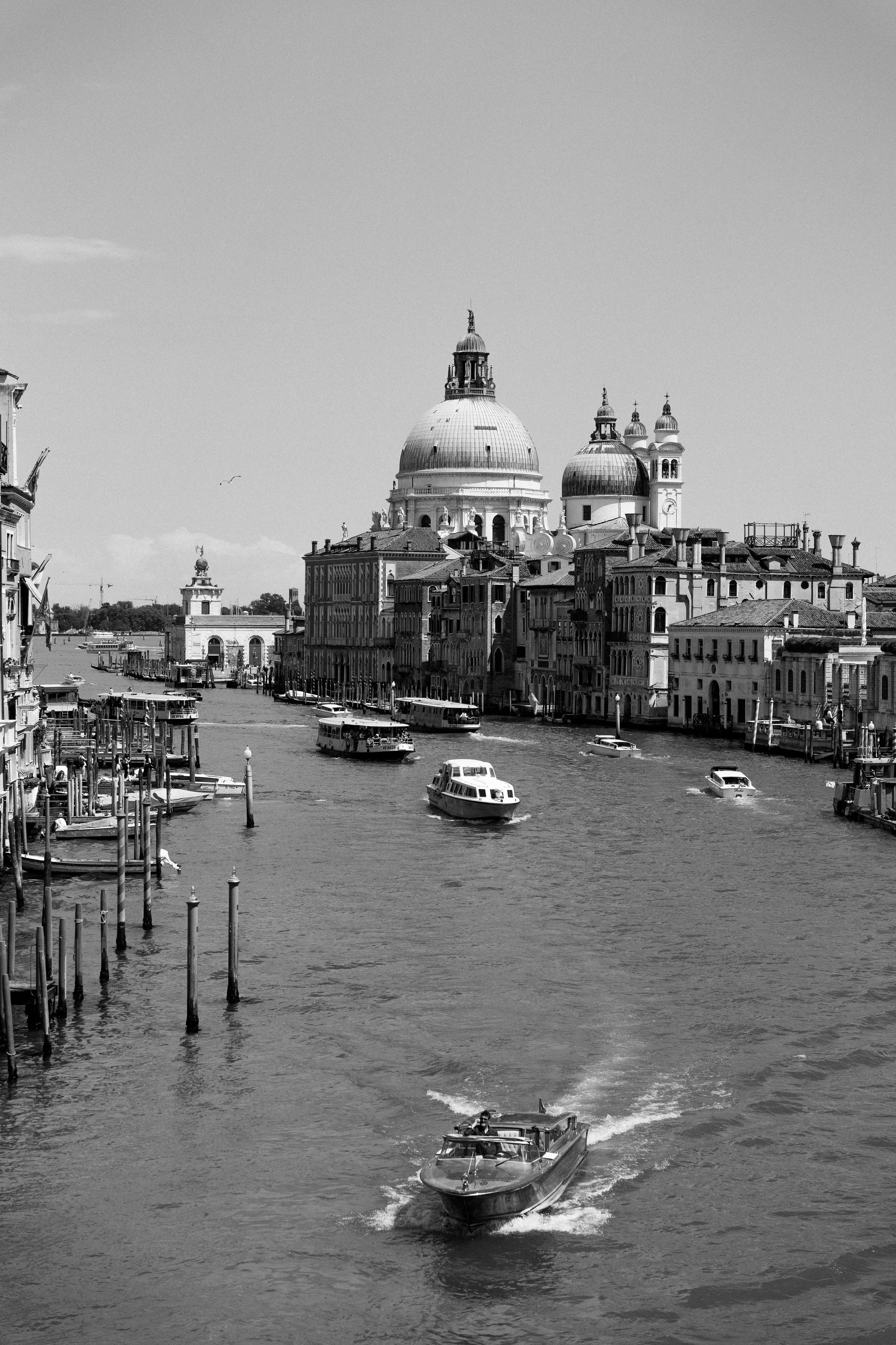 Black and white photo of a canal with boats and historic buildings, including domed churches, in Venice, Italy.