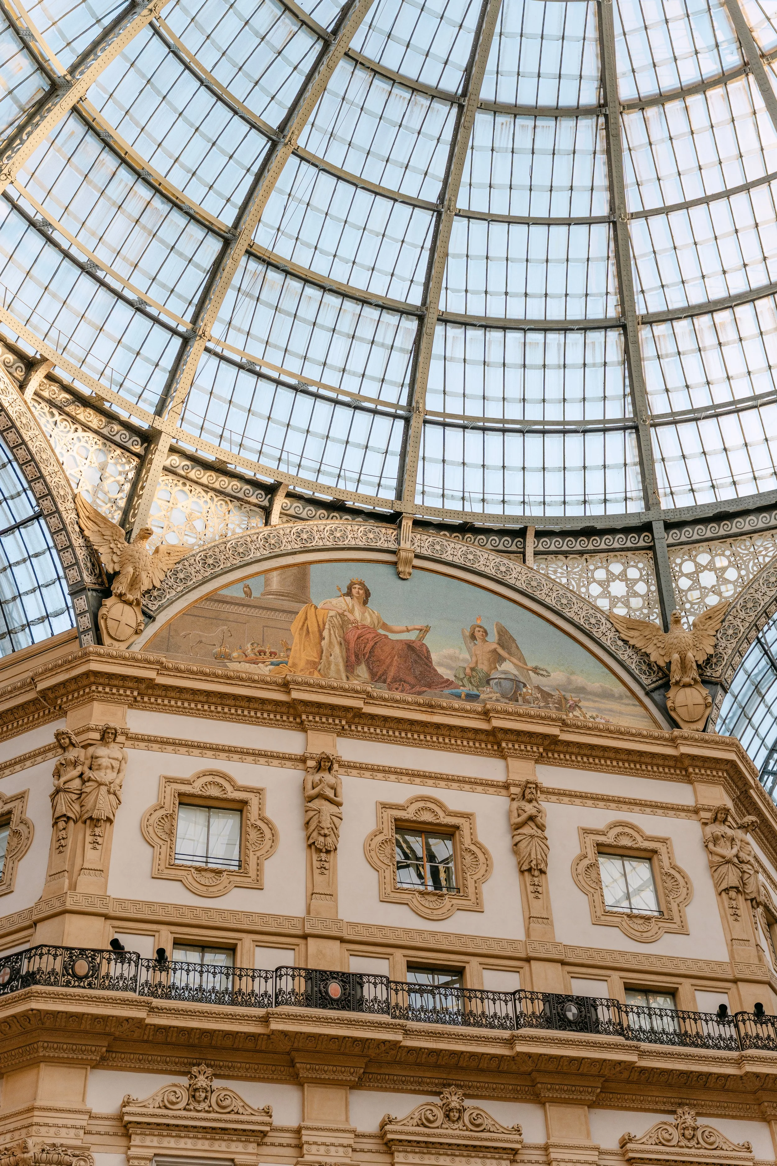 Interior view of a grand historic building with ornate architectural details, statues, and a painted mural, topped by a large glass dome ceiling.