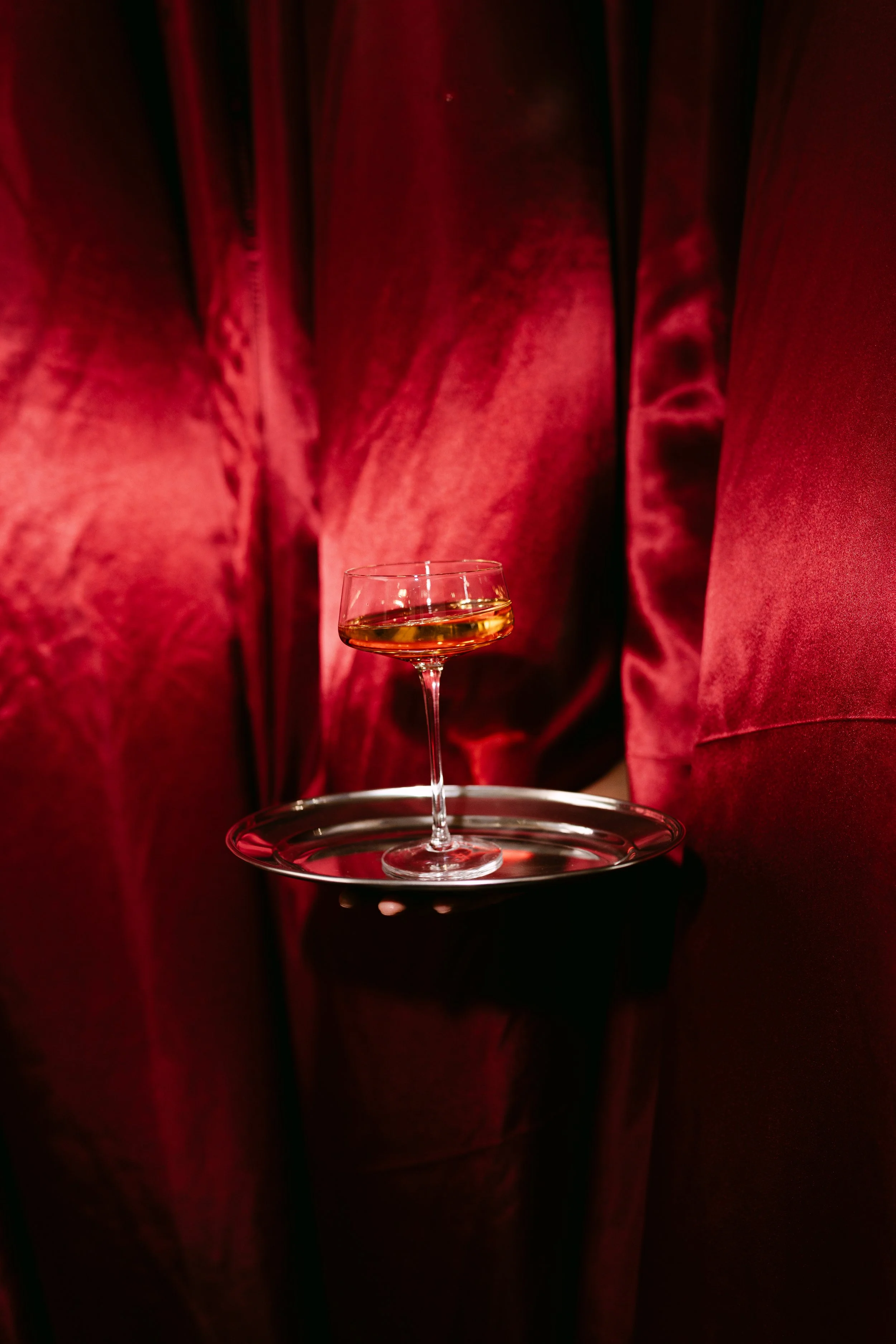 A glass of amber-colored wine on a tray held by a person in a red velvet outfit.