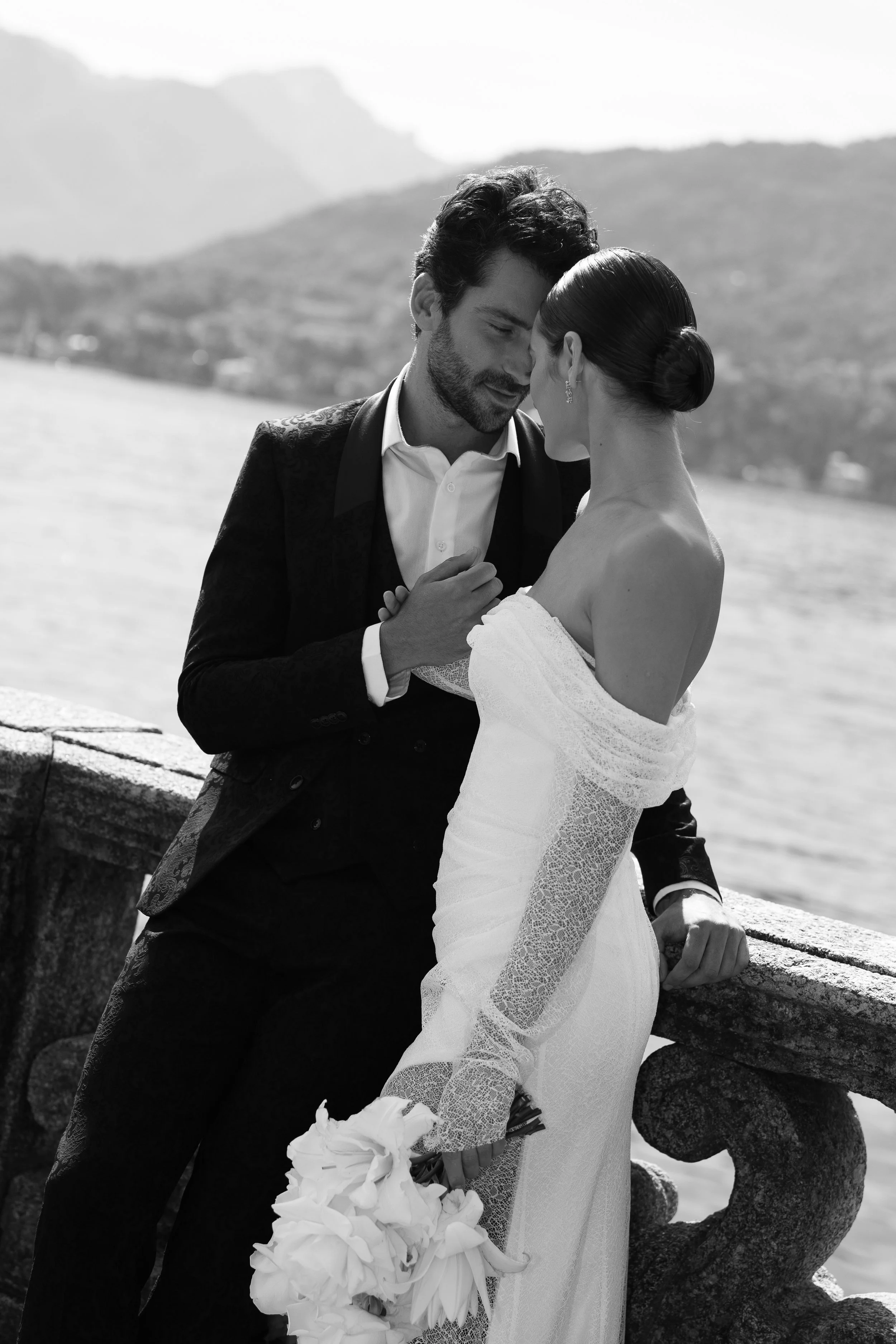 A black-and-white photo of a bride and groom embracing near a body of water with mountains in the background. The bride is holding a bouquet of flowers, and they are close with foreheads touching.