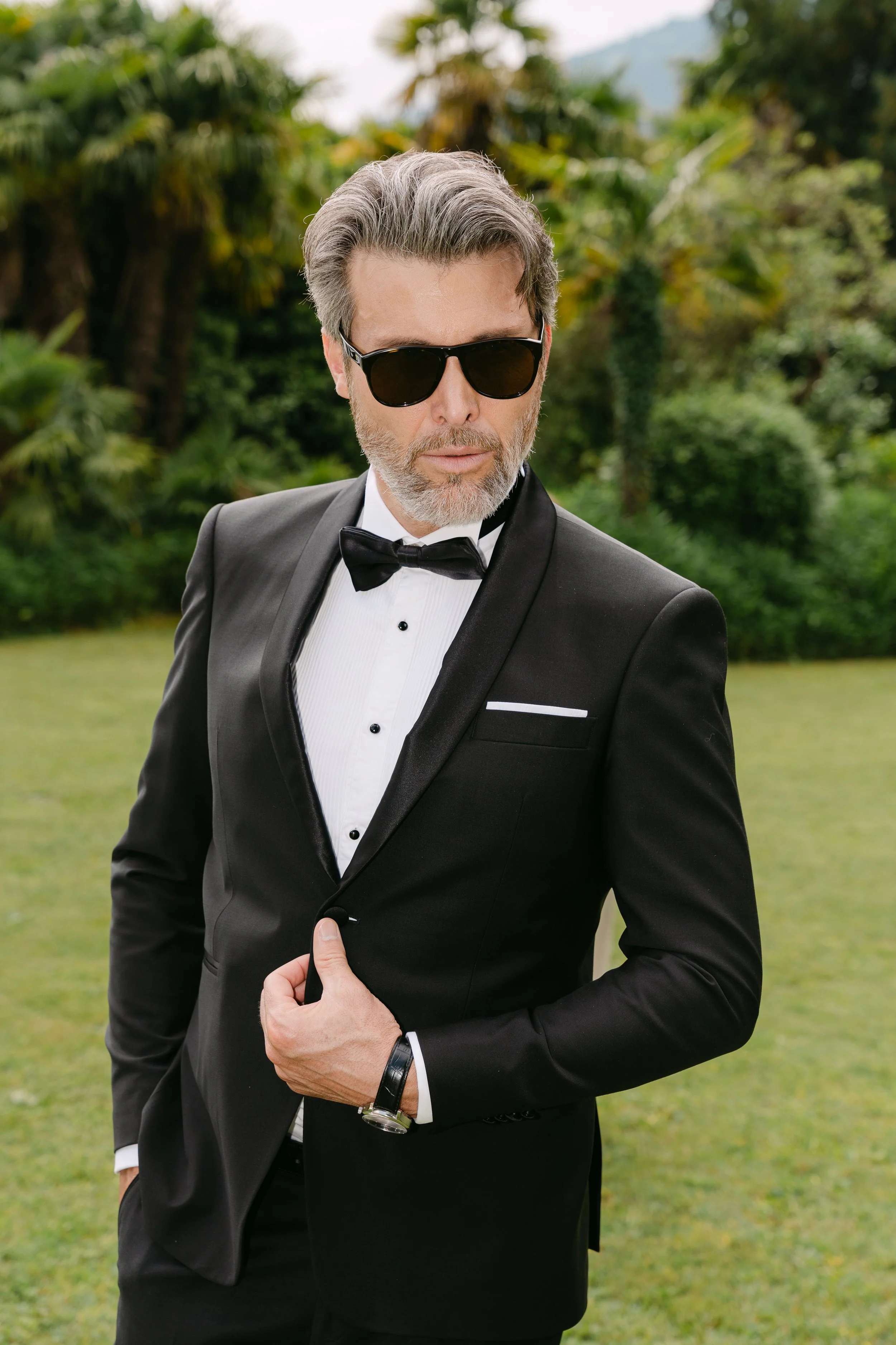 A man in a black tuxedo with a bow tie and sunglasses standing outdoors on a lawn with greenery and trees in the background.